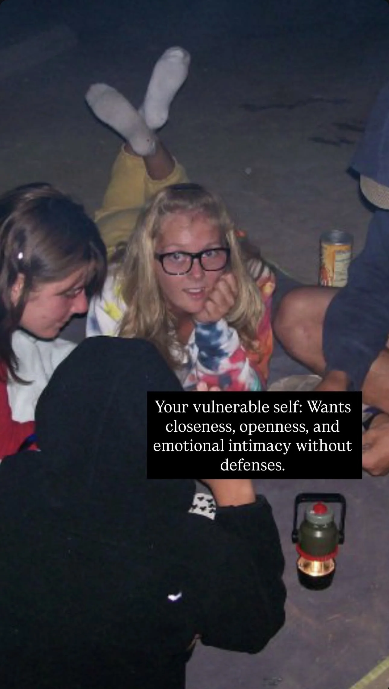 A group of people sitting on the ground at night, with one woman wearing glasses and resting her chin on her hand. There are snacks and a can on the ground, and a small camping lantern in the foreground. A black box with white text overlays the image