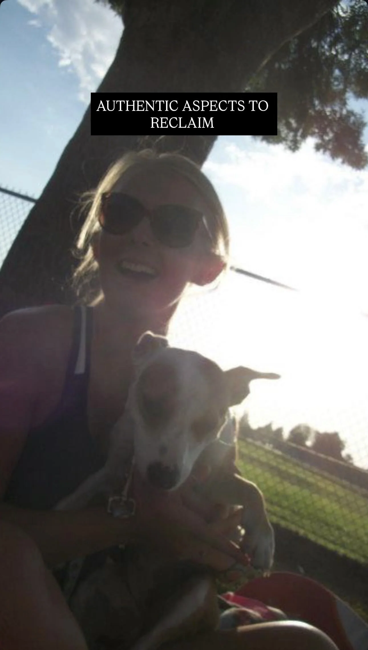 A woman wearing sunglasses holding a dog outdoors with a tree and a fence in the background, during daytime.