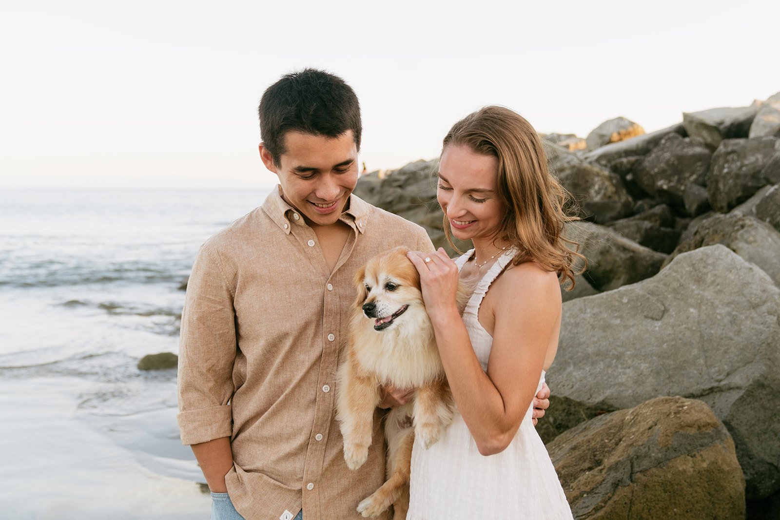 A smiling young couple holding a fluffy dog at the beach with rocks and the ocean in the background.