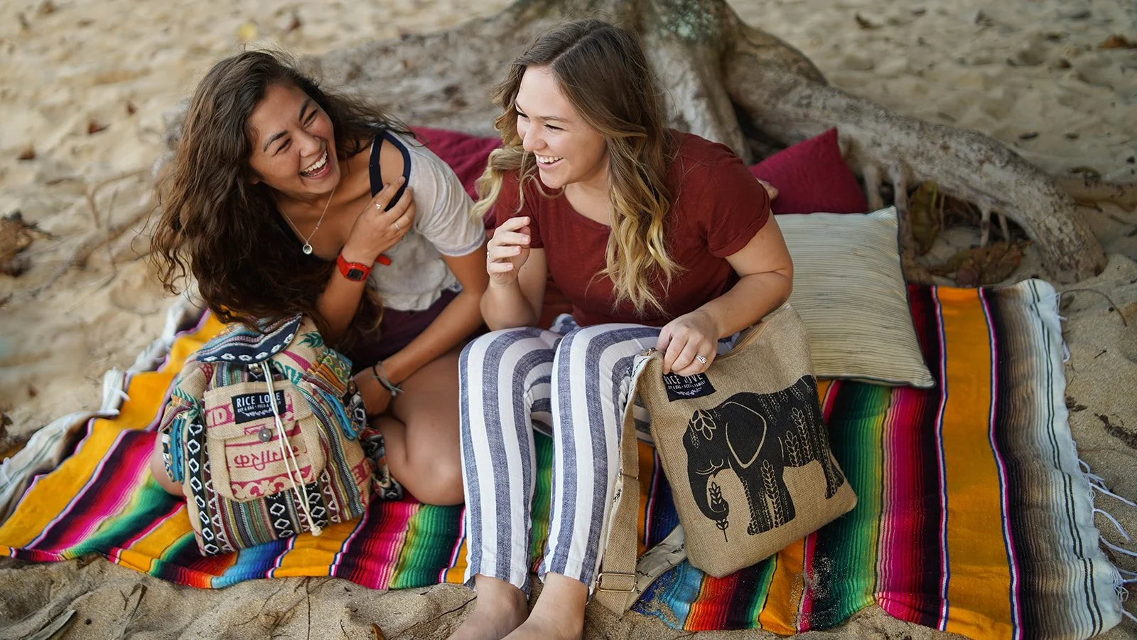 Two women sitting on a colorful blanket on the beach, laughing and talking, with pillows and a bag nearby.