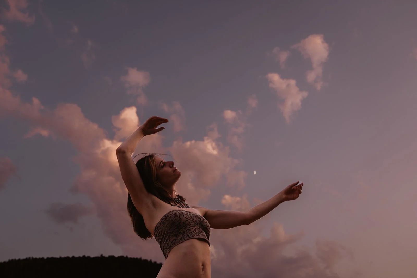Woman with long hair and a strapless top standing outdoors at sunset or dusk, reaching up towards the sky with clouds and a crescent moon in the background.