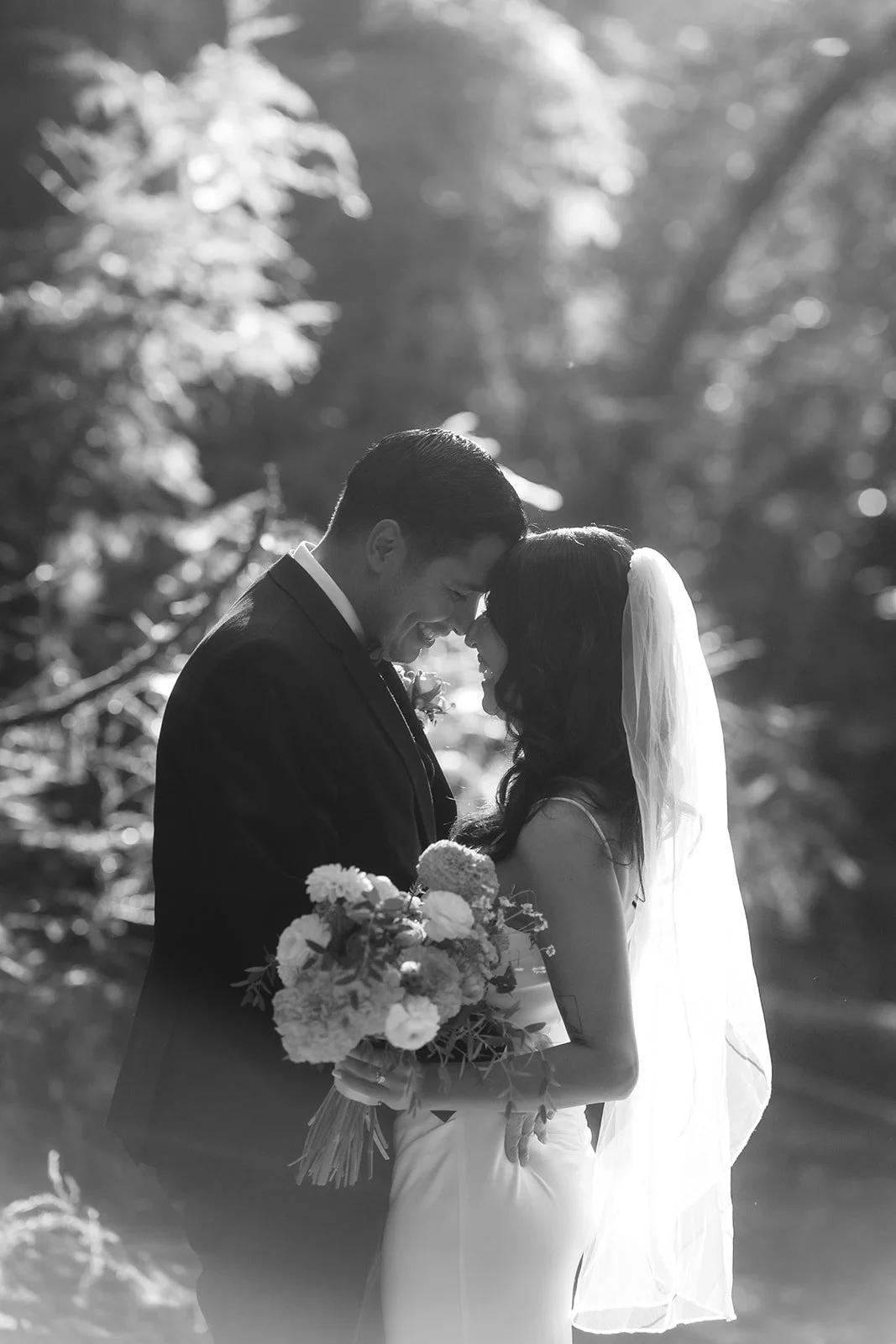 A bride and groom facing each other, touching foreheads, outdoors with sunlight filtering through trees, the bride holding a bouquet of flowers.
