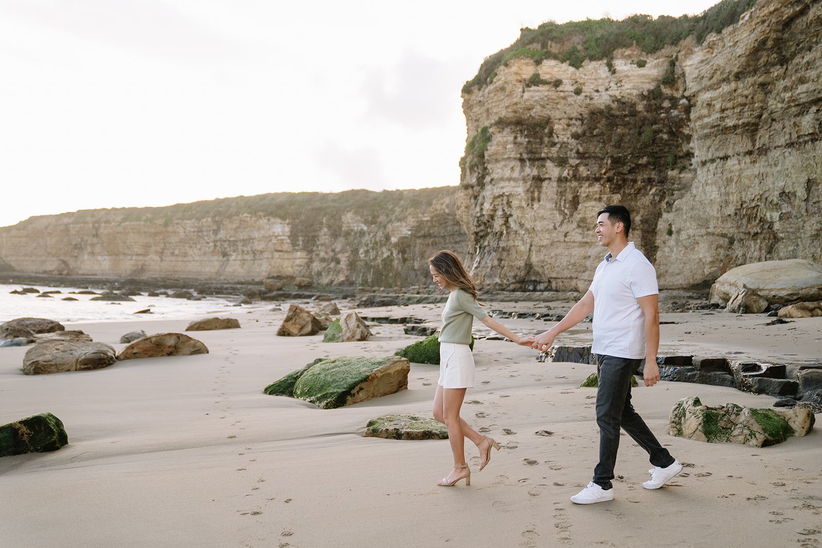 A couple walking hand in hand on a sandy beach with rocks and cliffs in the background during sunset.