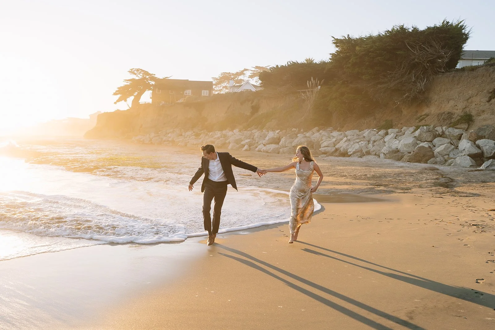 A couple dressed in formal clothing walking hand in hand on a sandy beach during sunset, with ocean waves and coastal houses in the background.
