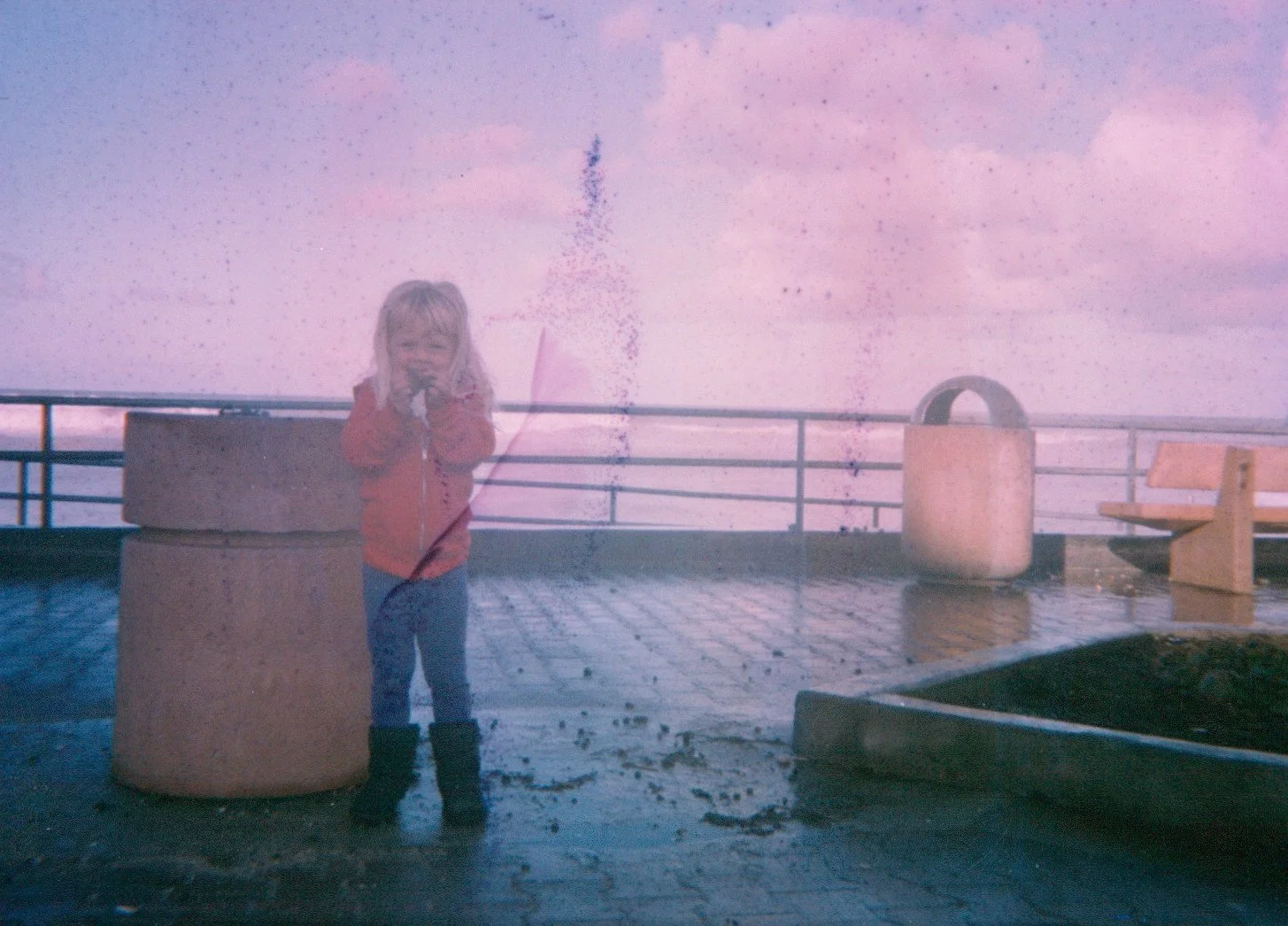 A girl in a red jacket and boots standing on a wet pier, holding a stick with pink ribbons, with a pink sky and water in the background.