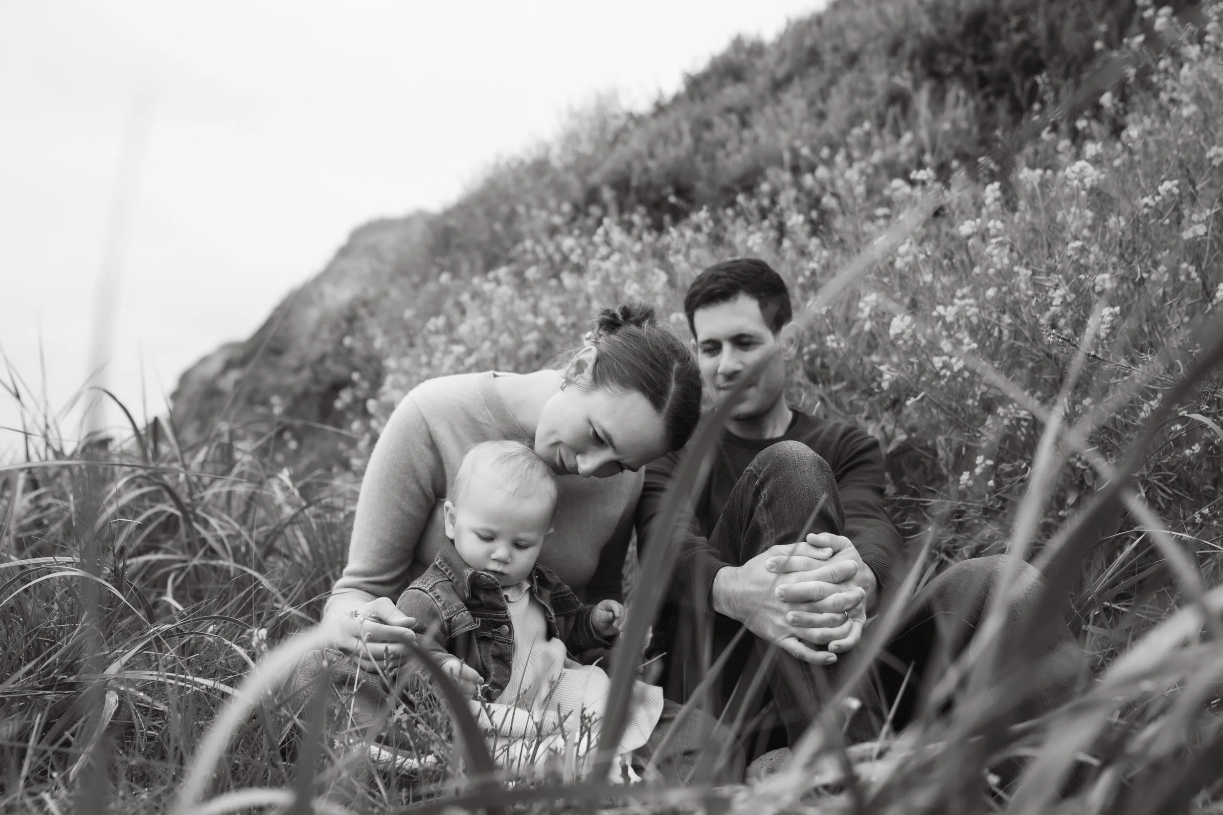Black and white photo of a happy family consisting of a woman, a man, and a young child sitting outdoors in a grassy area with a hilly landscape in the background.