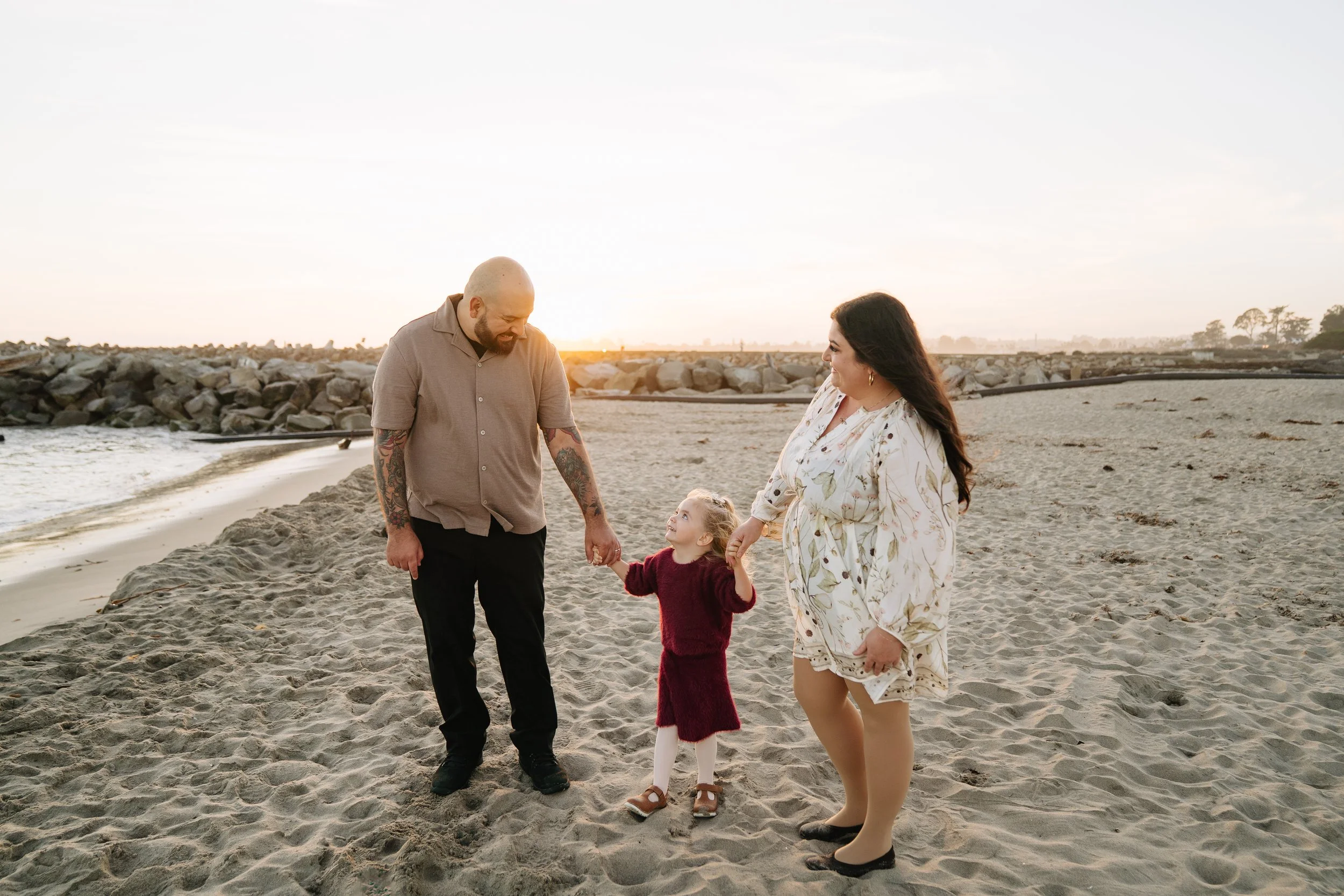 A family of three, a man, a woman, and a young girl, holding hands and walking on a sandy beach during sunset.
