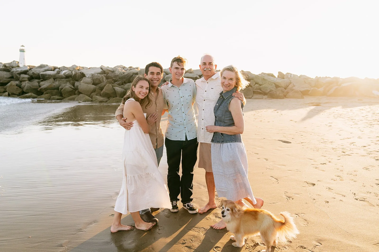 Family of five with two men, two women, and a teenage boy, standing on a sandy beach with a small dog, rocks, and a lighthouse in the background, during sunset.