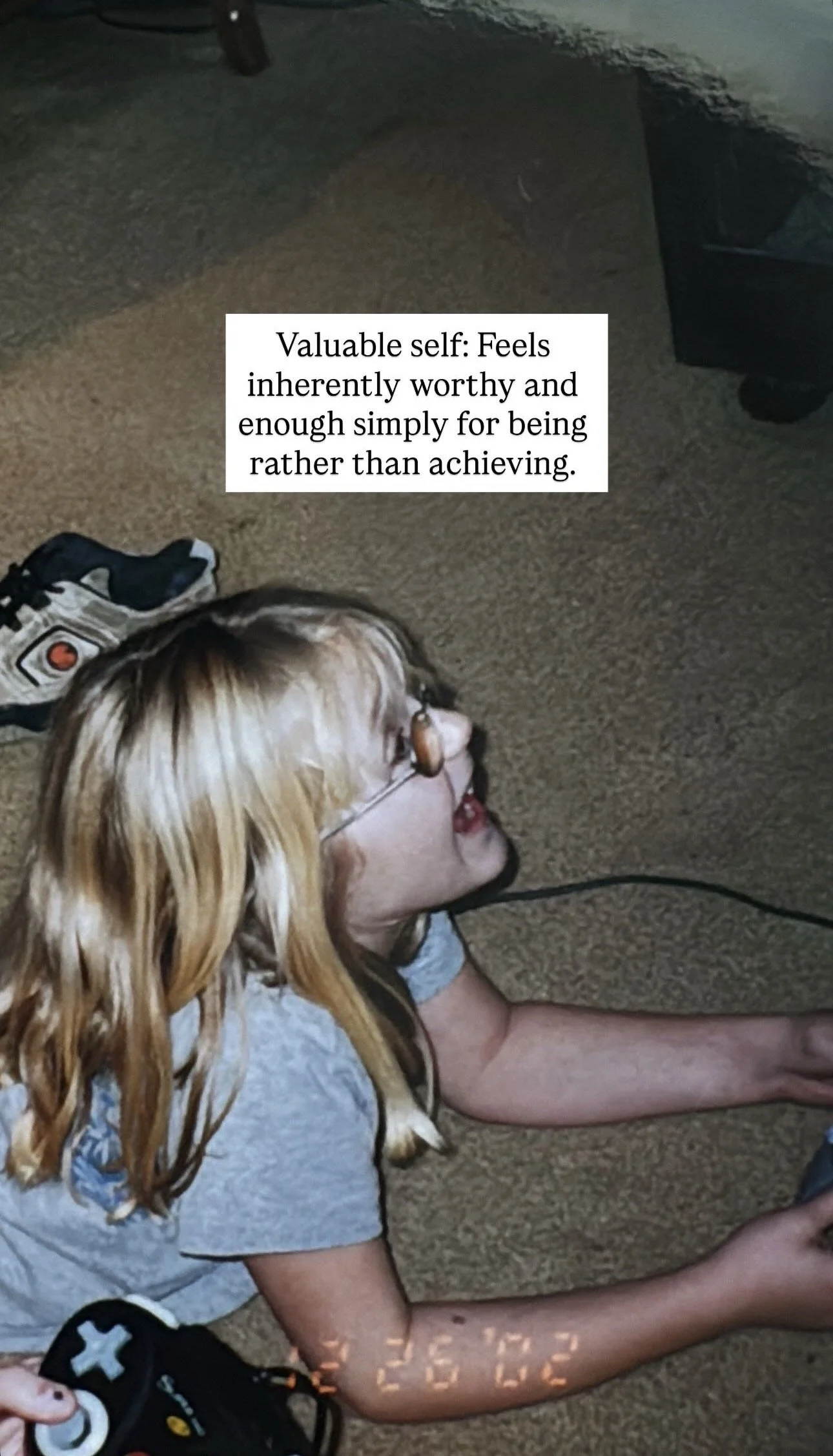 A young girl with blonde hair lying on her stomach on a carpeted floor, wearing glasses and a gray shirt, with a game controller near her hand. A motivational quote about self-worth is overlaid on the image.