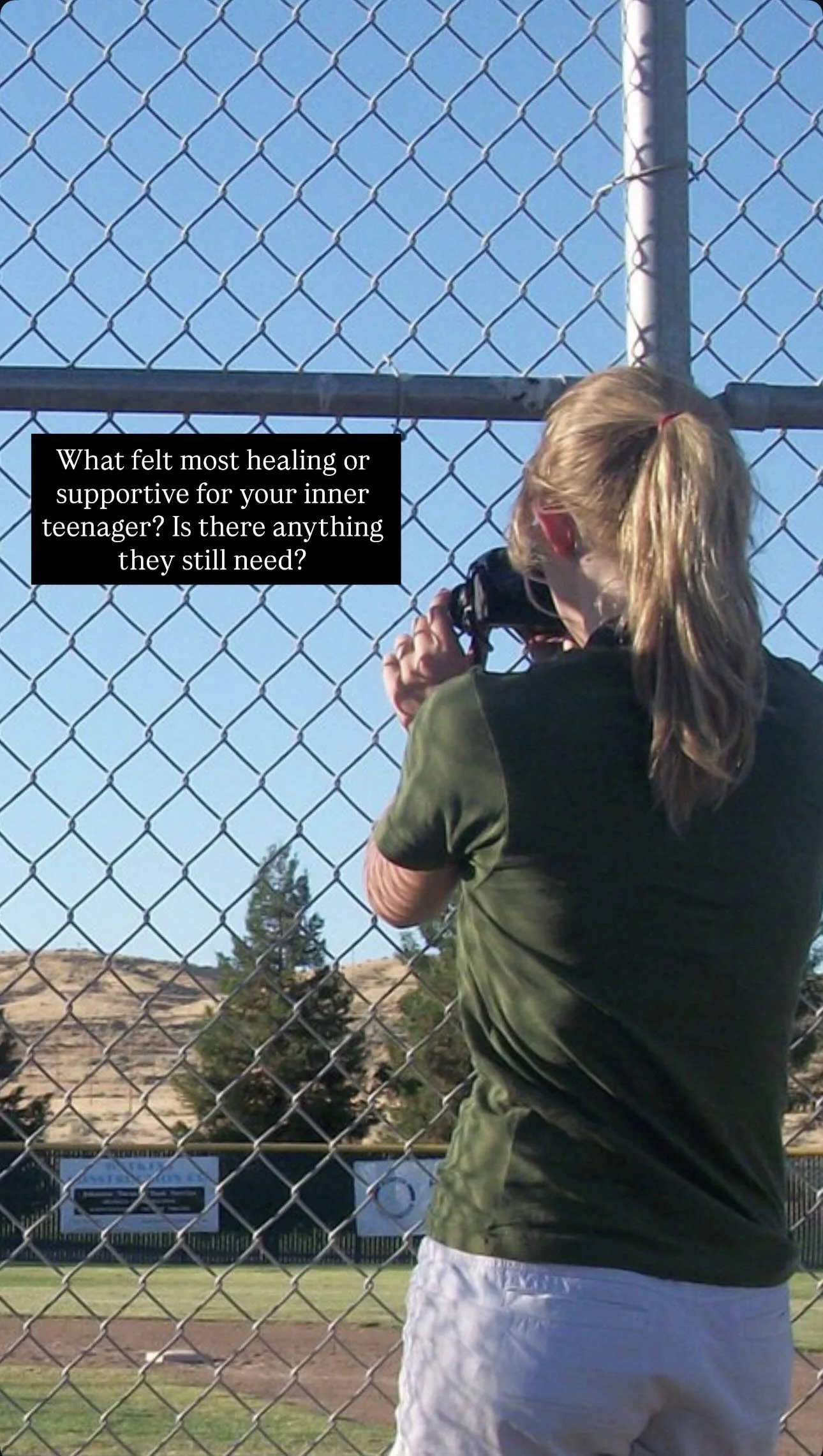 A young girl with blonde hair in a ponytail, wearing a green shirt and white shorts, stands behind a chain-link fence at a baseball field, holding a camera while watching the game. The sky is clear and blue, with some trees and a hillside in the back