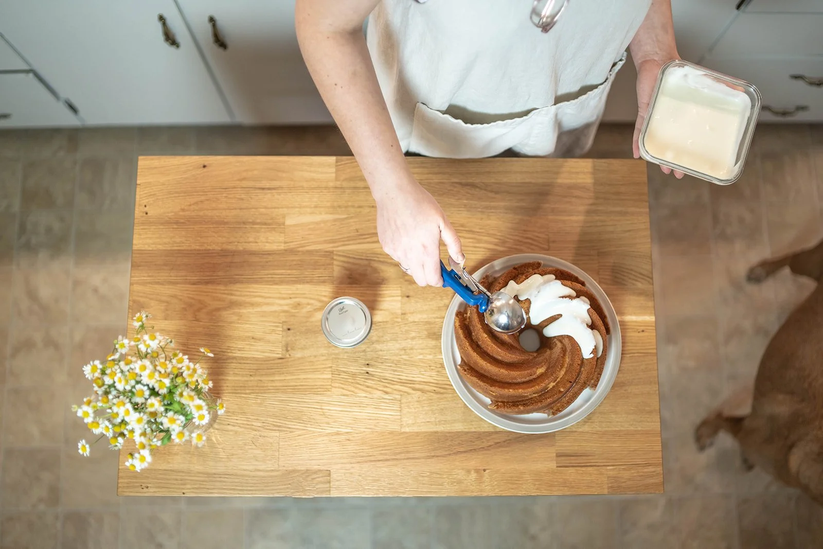 A person wearing a white apron is adding whipped cream to a Bundt cake on a wooden table. The person is holding a container of whipped cream in their left hand and a spoon in their right hand. There is a small vase of daisies, a jar lid, and a can of whipped cream on the table.