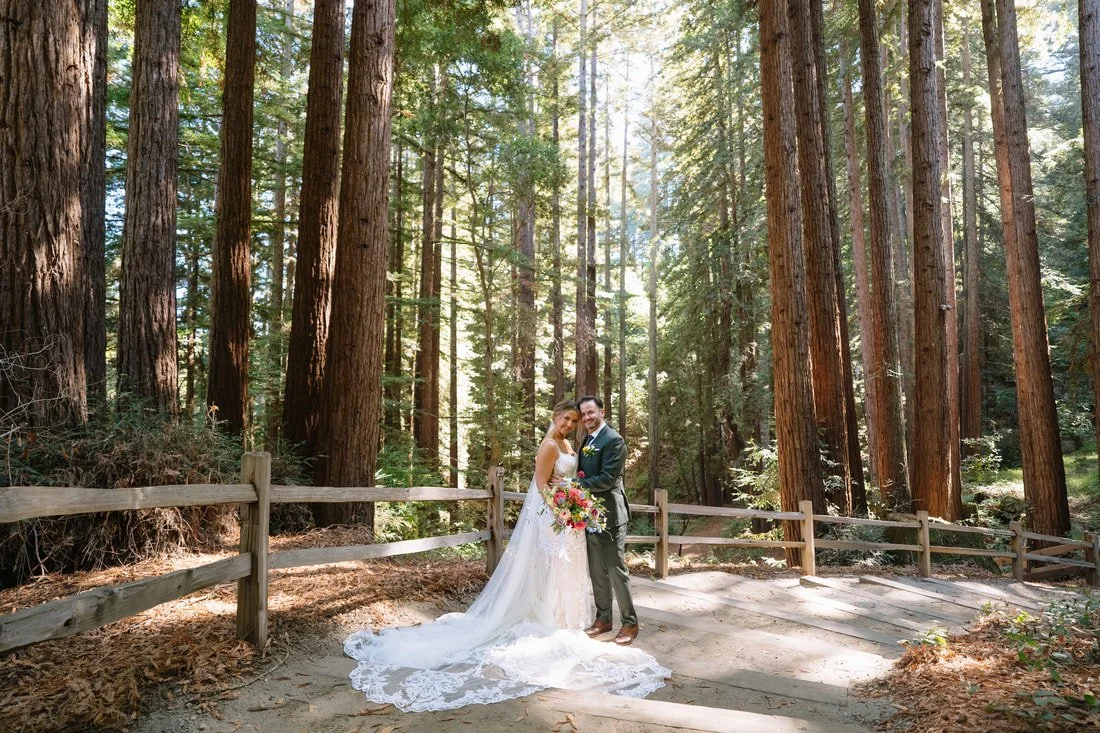 Bride and groom standing on a dirt forest trail surrounded by tall trees, holding a bouquet, and smiling at the camera.