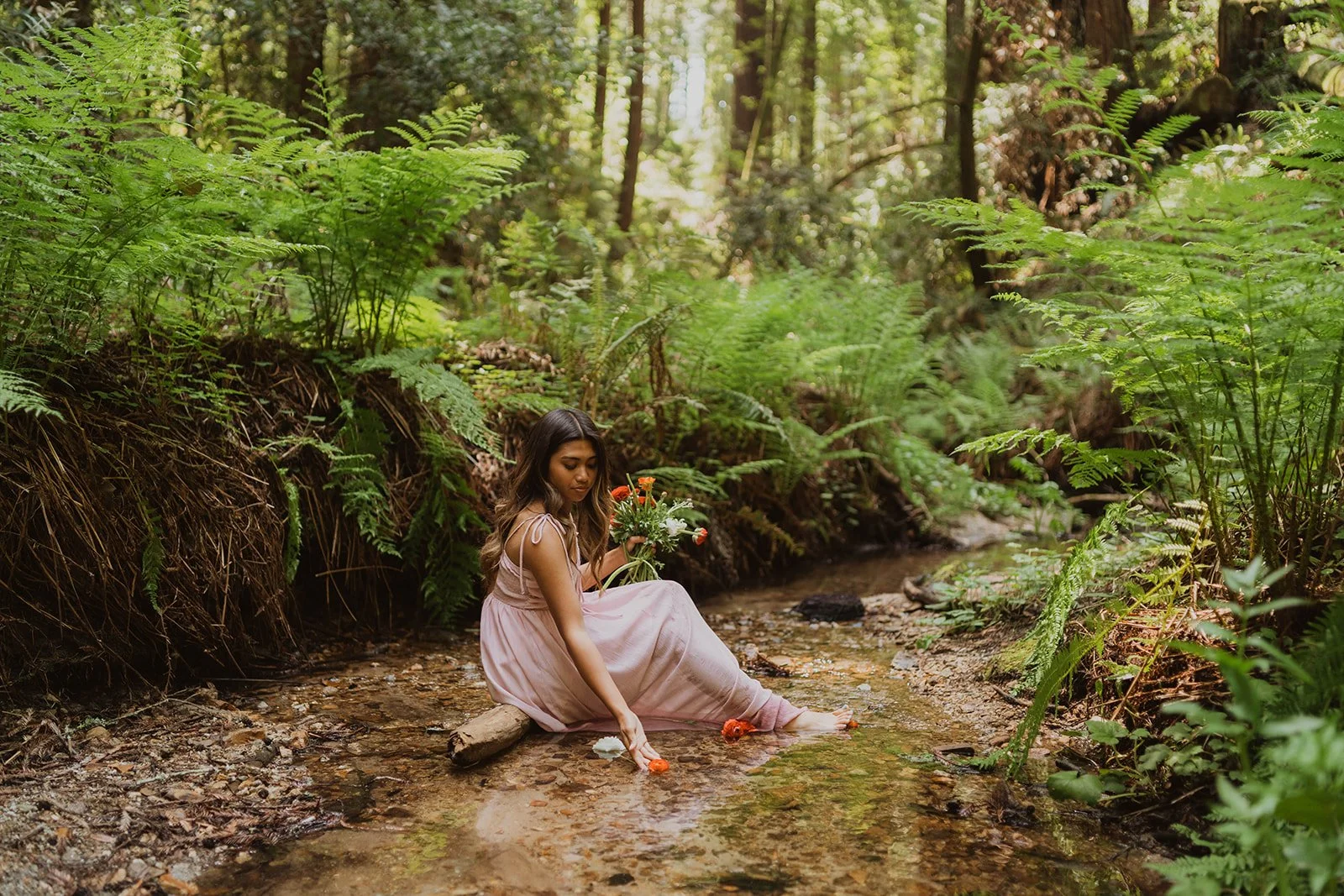 A woman in a pink dress sits on a log by a creek in a lush, green forest, holding a bouquet of orange and white flowers and touching the water with her hand.