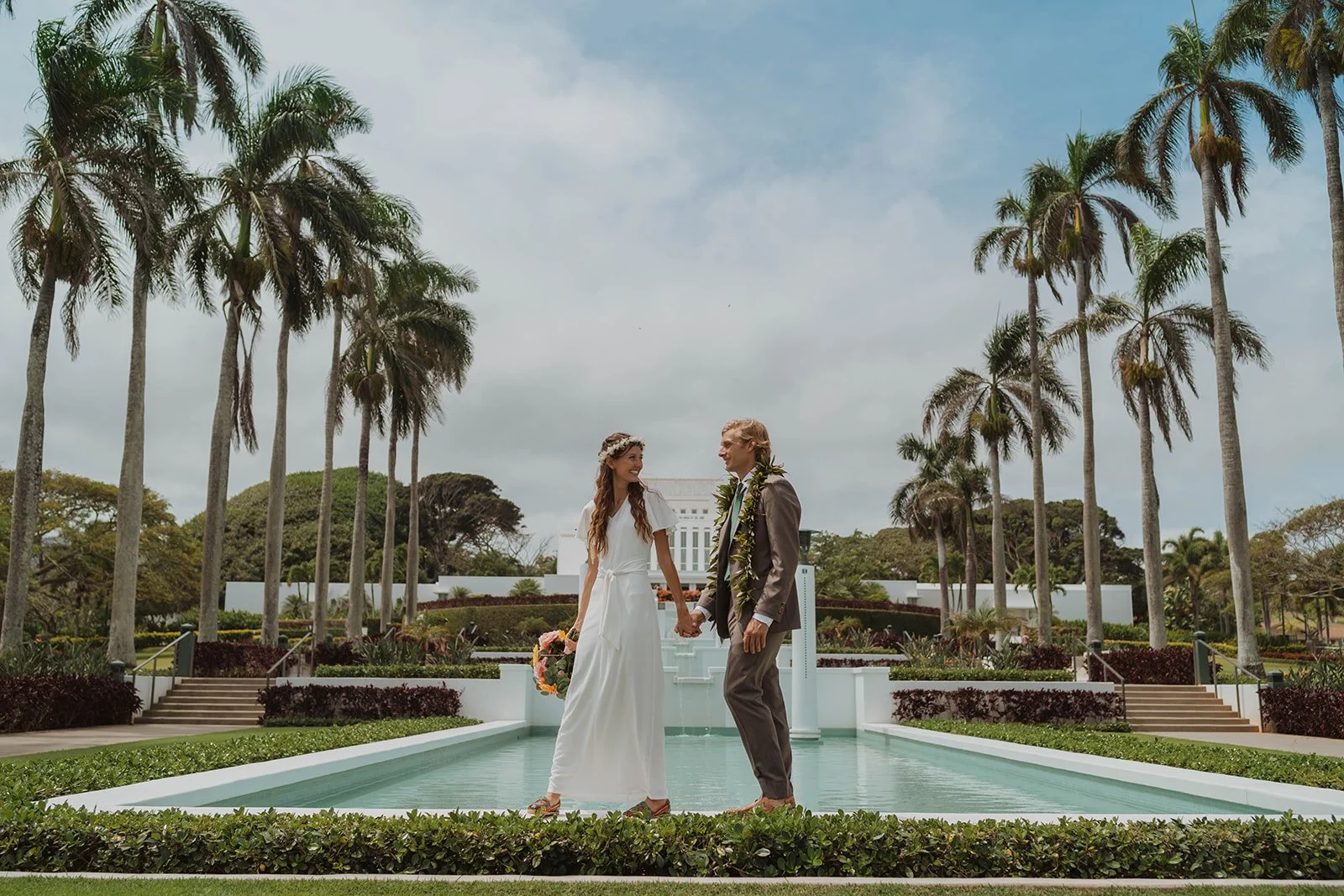 A bride and groom holding hands and standing in a pool in a lush garden with tall palm trees during a wedding ceremony.