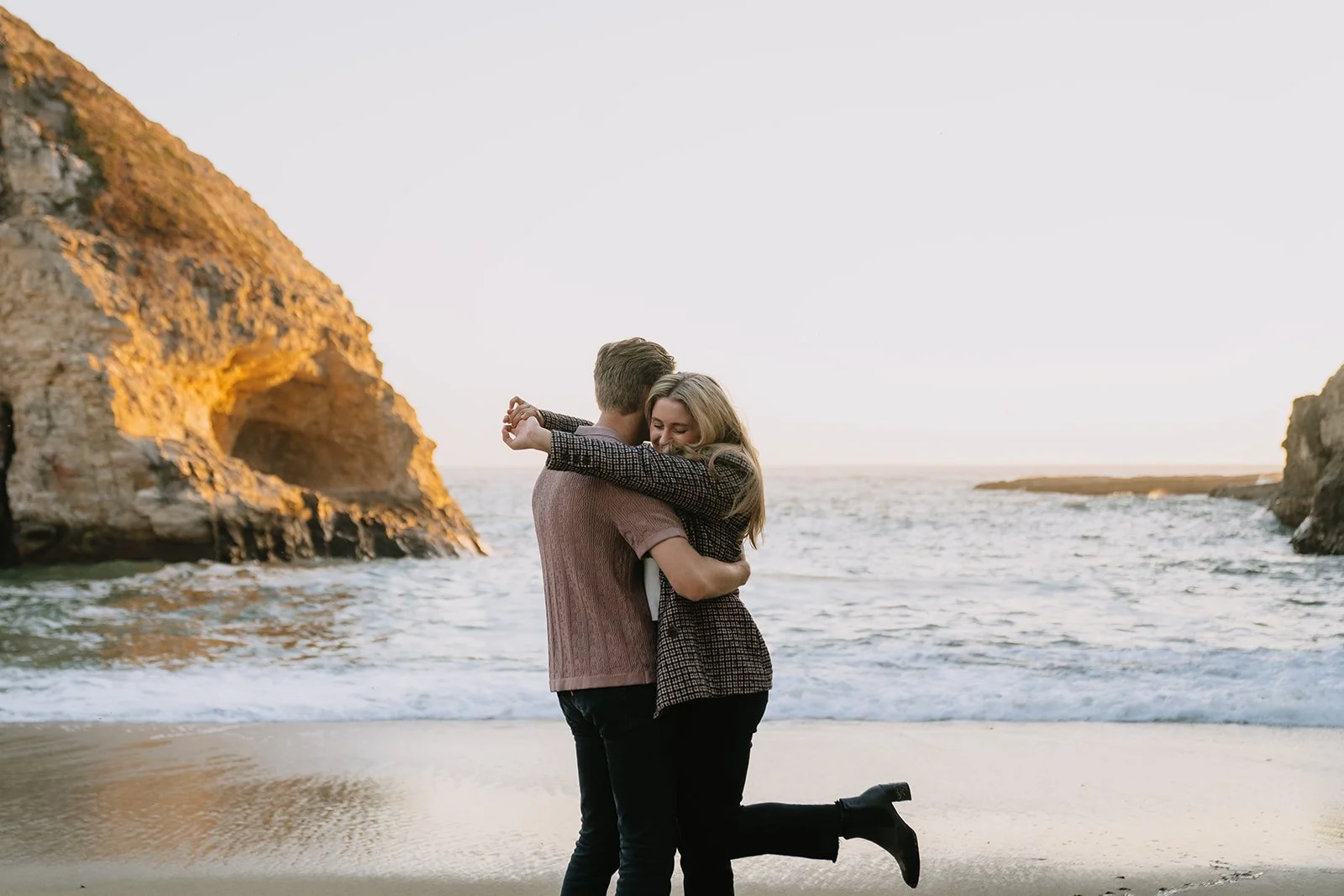 A couple hugging on a beach during sunset, surrounded by cliffs and ocean.