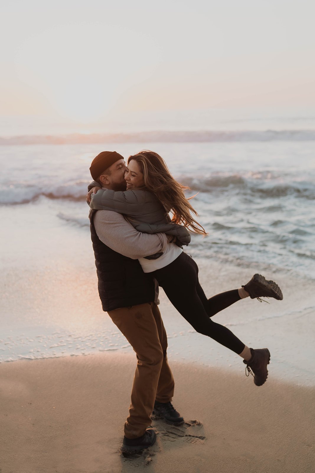 A couple hugging on the beach at sunset, with ocean waves in the background.