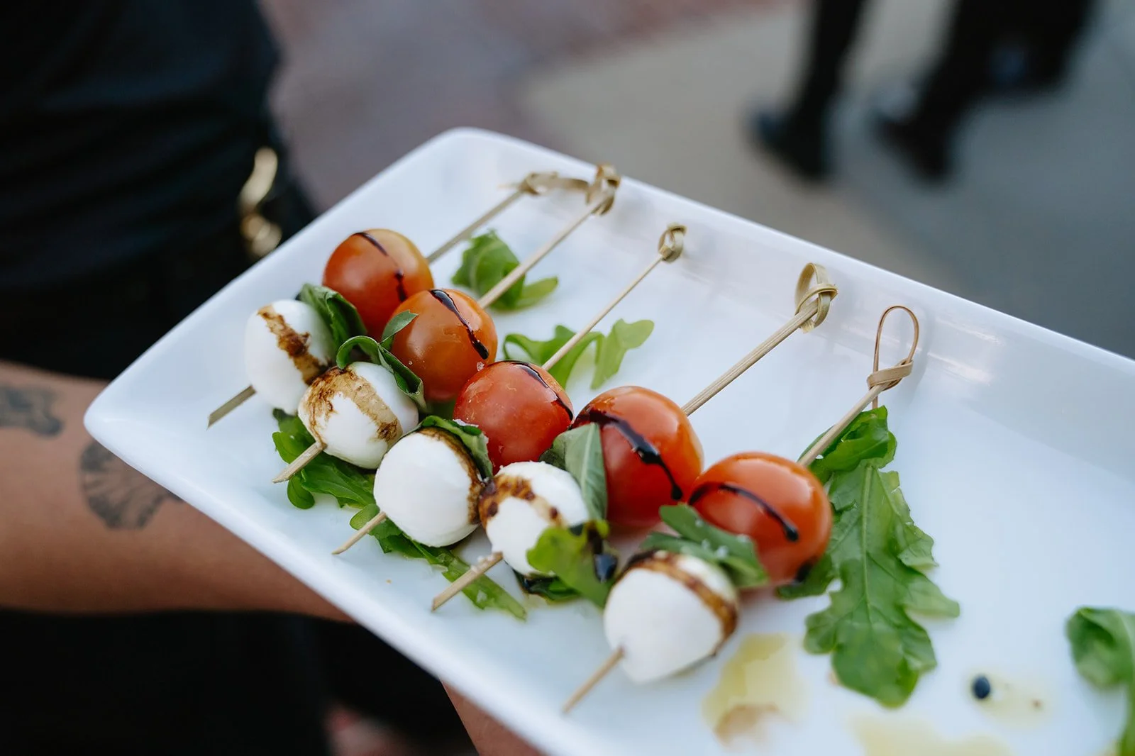 A white rectangular plate holding skewers of cherry tomatoes, mozzarella balls, and basil leaves, drizzled with balsamic glaze, on a bed of arugula leaves.