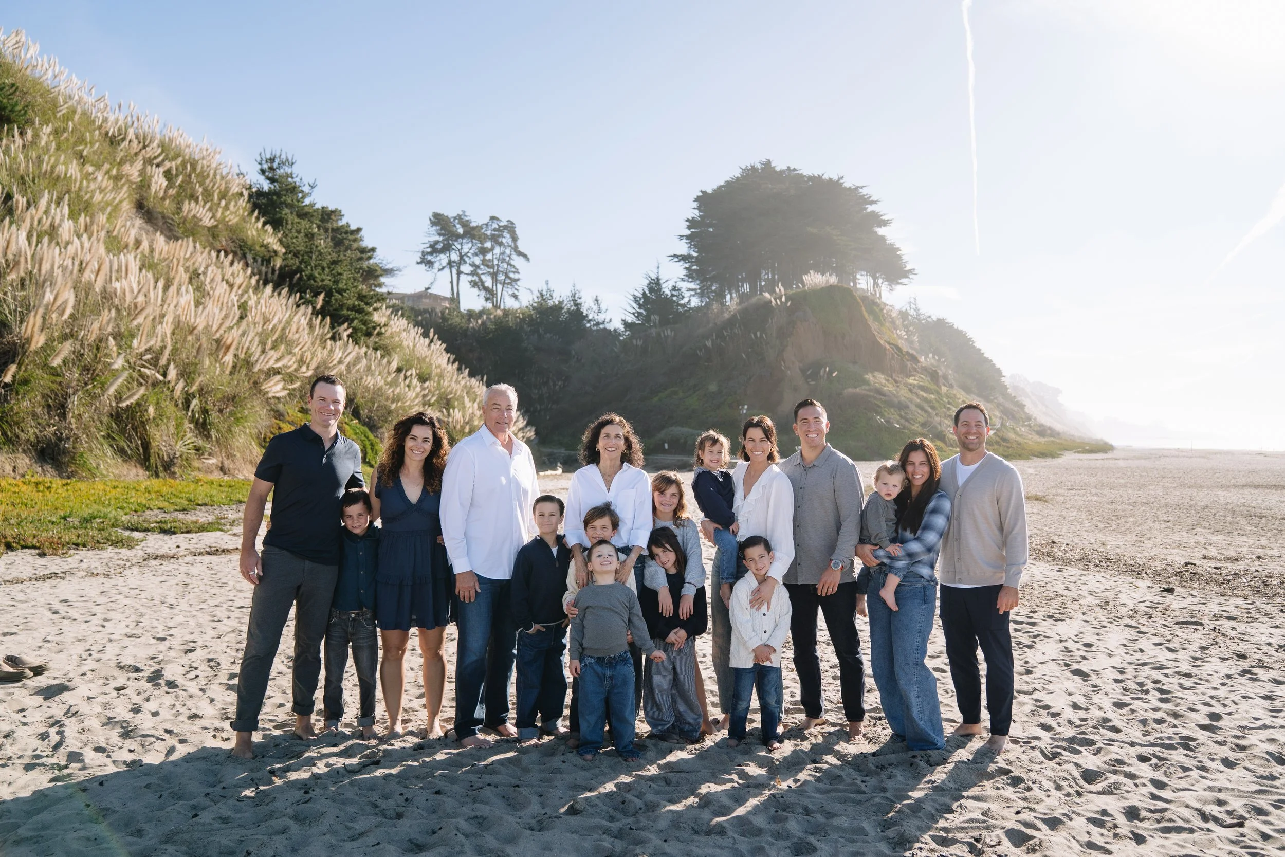 Family with children standing on the beach near a hillside with tall grass and trees under a clear sky.