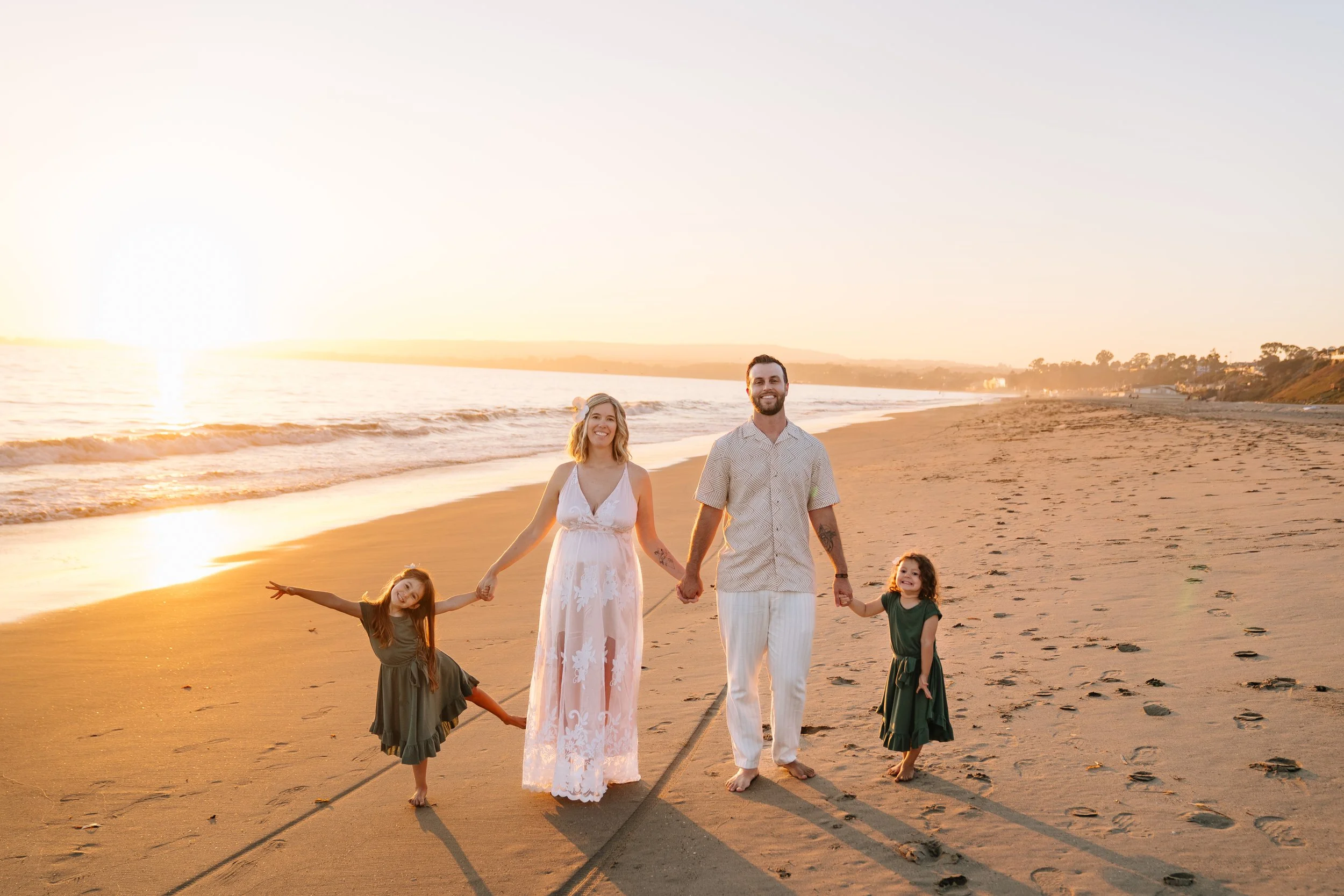 A family of four walking on the beach at sunset, holding hands and smiling. The parents are in the center with two young girls on either side, all dressed in summer clothes.