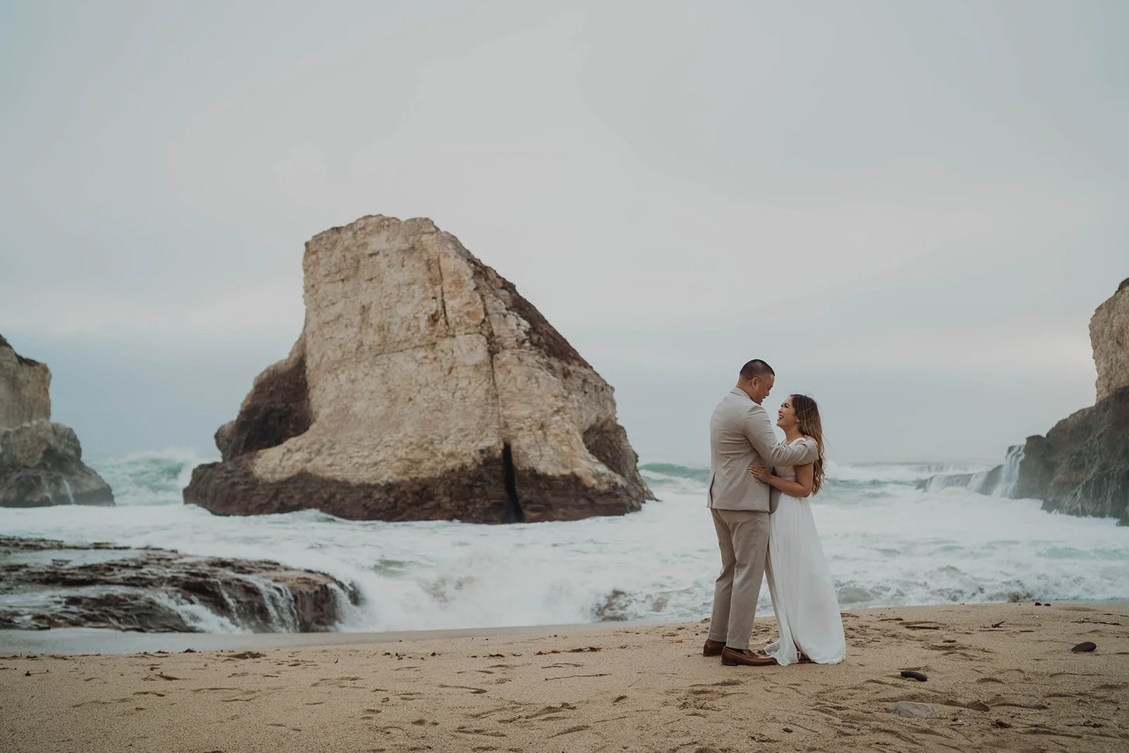 A couple in wedding attire standing on a beach with large rocks and waves in the background.