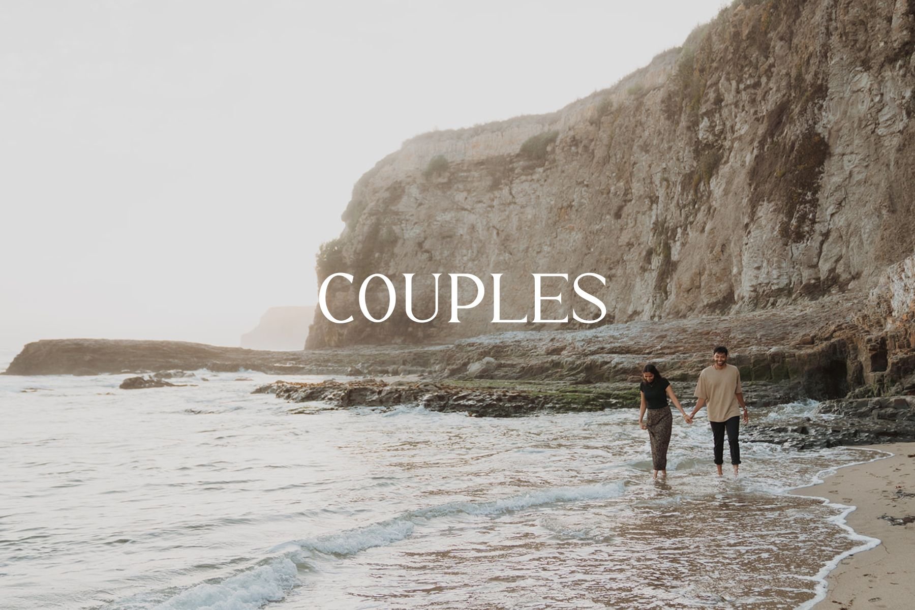 A couple walking hand in hand along the beach shoreline with cliffs in the background.