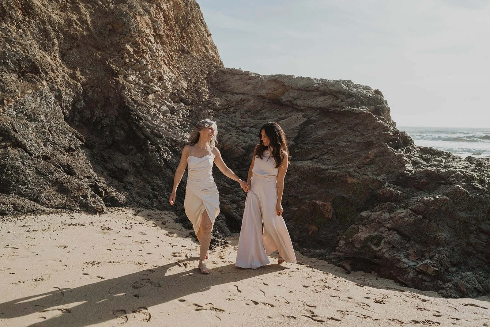 Two women in white dresses walking hand in hand on a sandy beach near rocky cliffs, smiling and enjoying the sunny day.