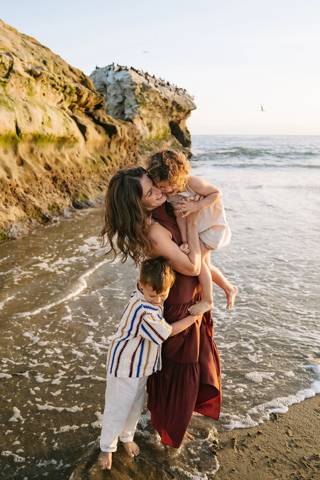 A woman with two children on a beach during sunset, hugging and playing near the shoreline with large rock formations and birds flying in the background.