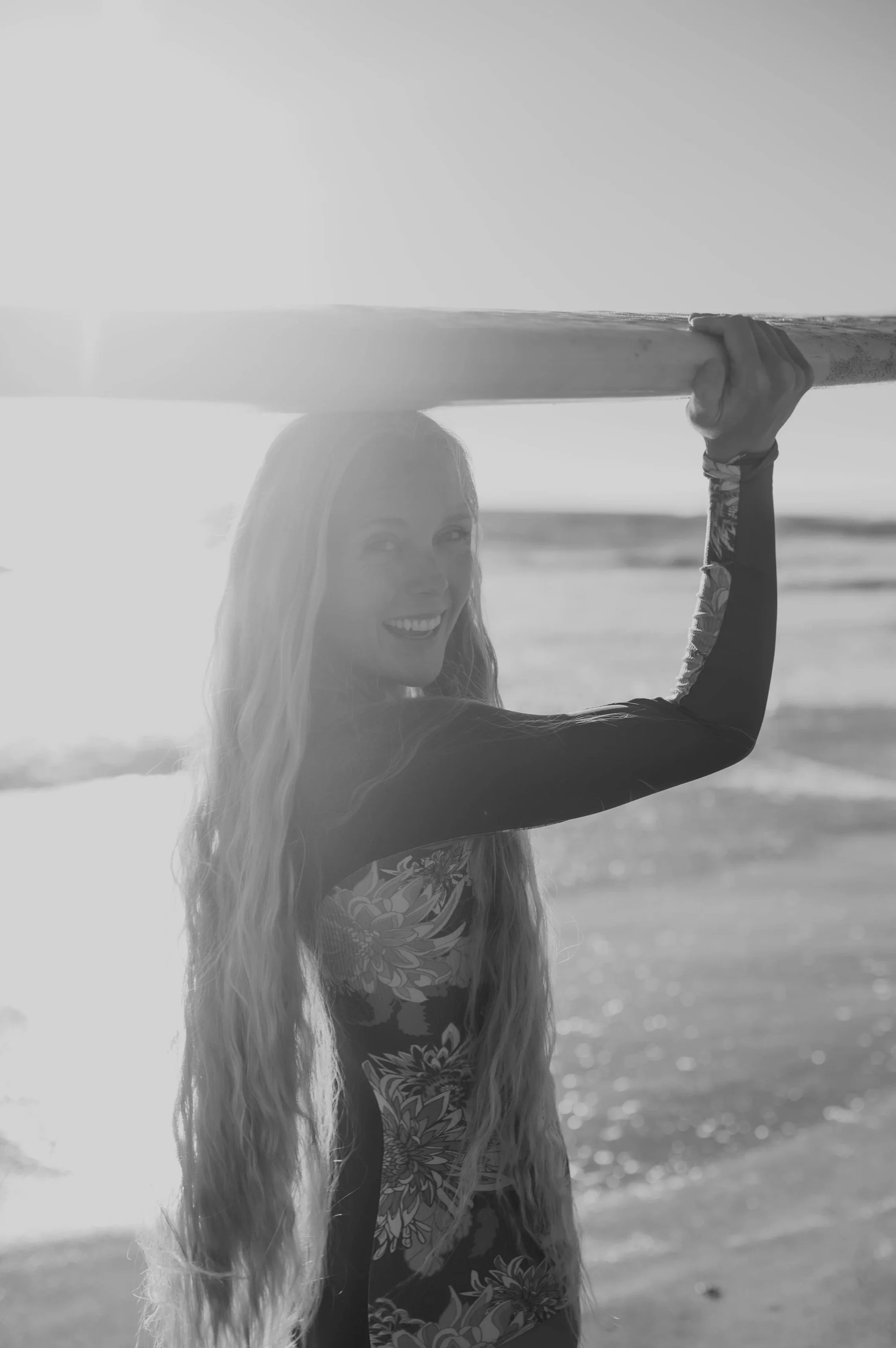Woman with long hair holding surfboard on beach at sunset, smiling at camera in black and white.