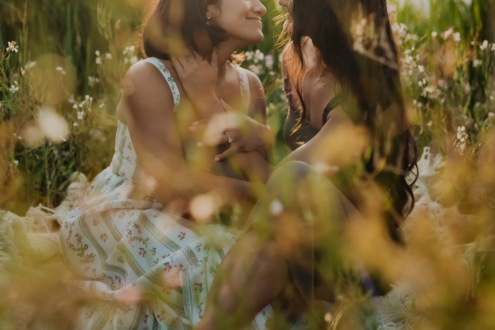 Two women sitting close together in a field of flowers, smiling and holding hands, surrounded by greenery and flowers.
