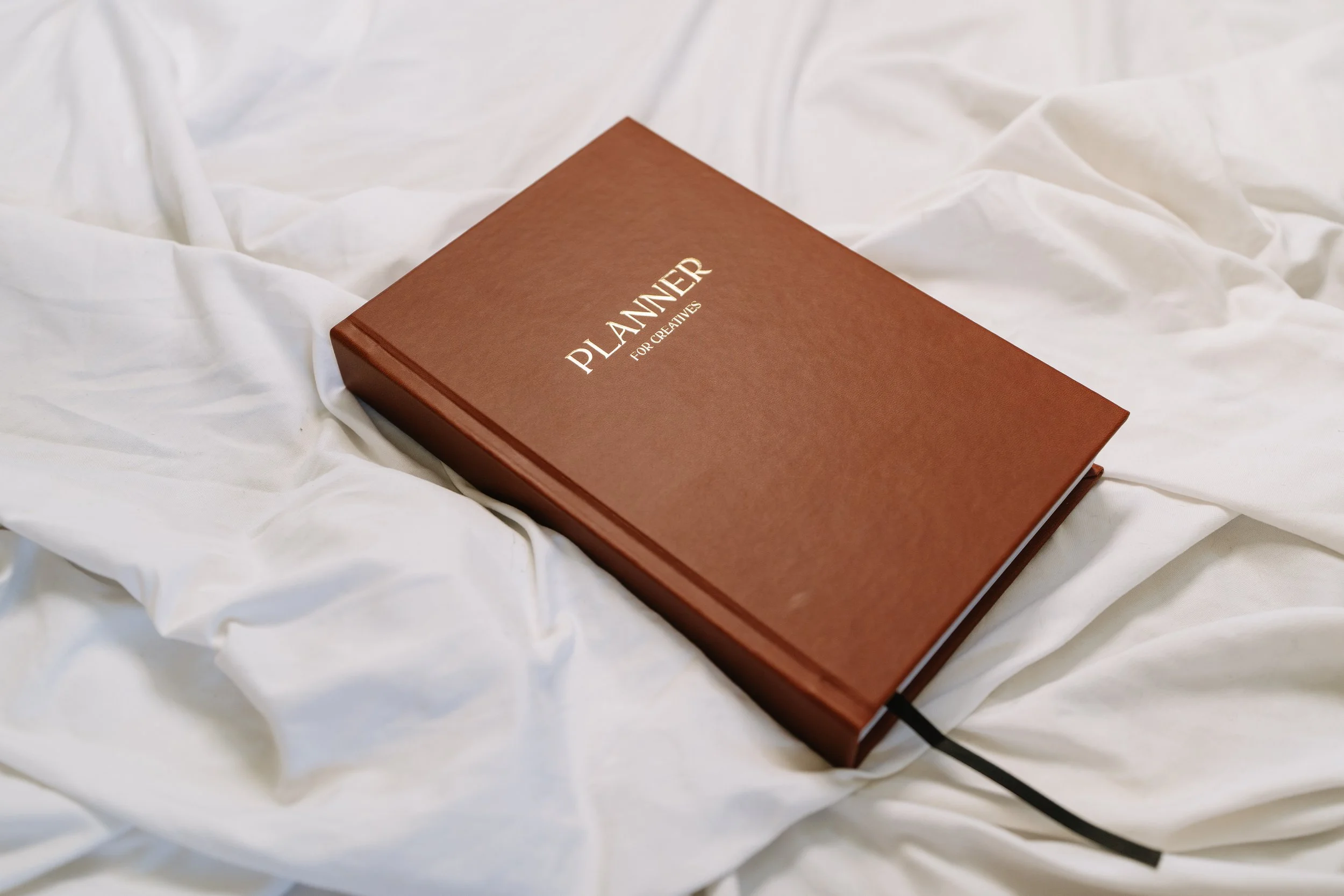 A closed brown planner with gold lettering on the cover that reads 'PLANNER FOR CREATIVES,' resting on white fabric.