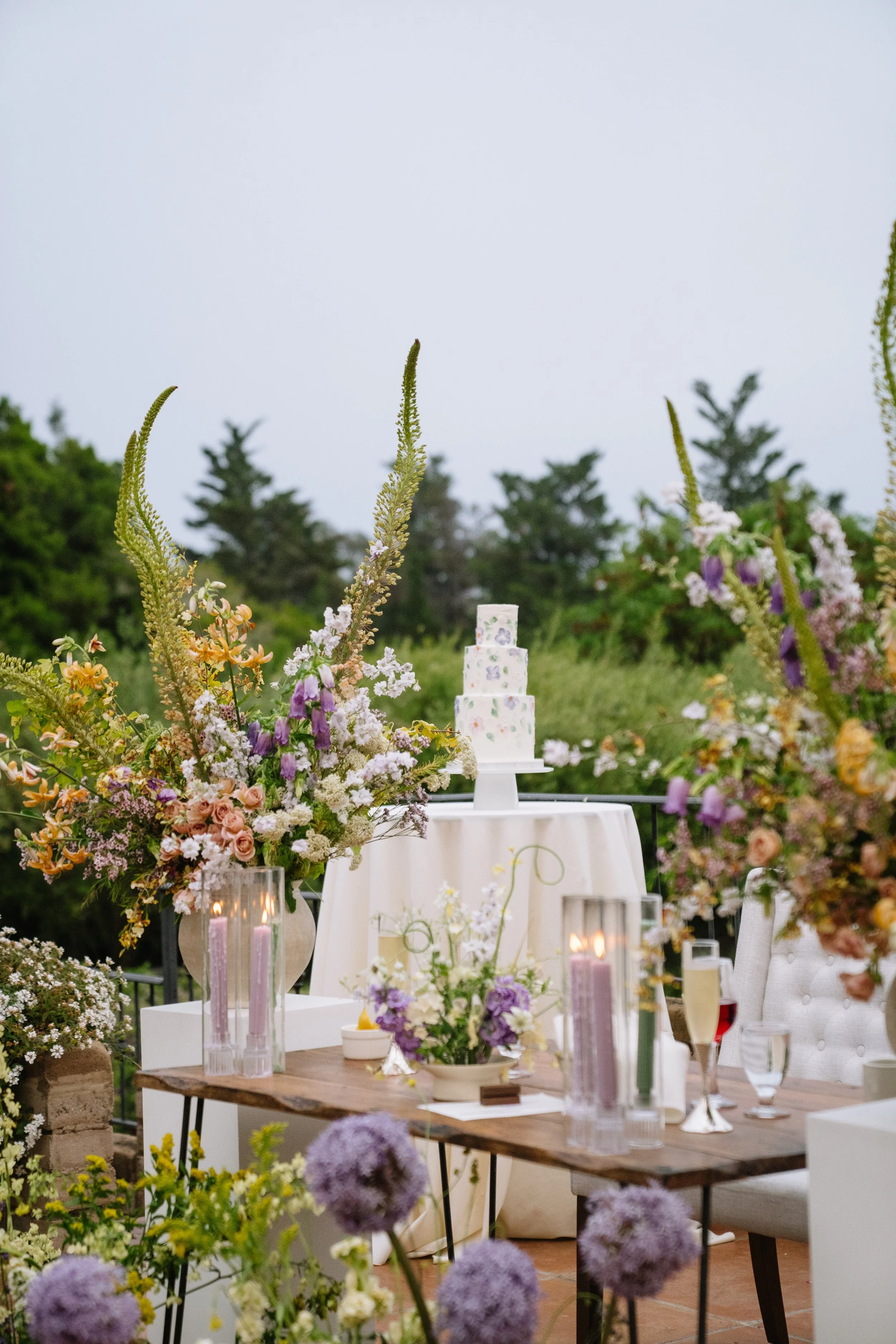 Outdoor wedding reception setup with a cake on a stand, floral arrangements, candles, and glasses on a table surrounded by greenery and flowers.