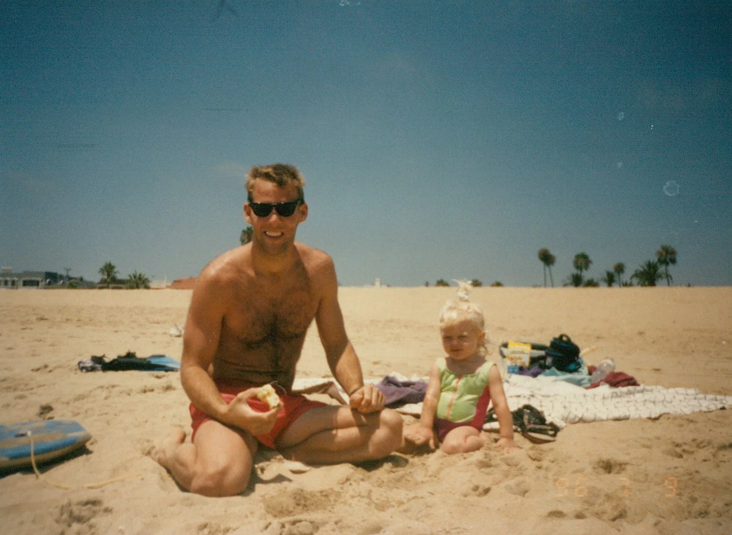 A man and a young girl sitting on the sandy beach during daytime, with towels and belongings scattered around, and palm trees in the background under a clear blue sky.
