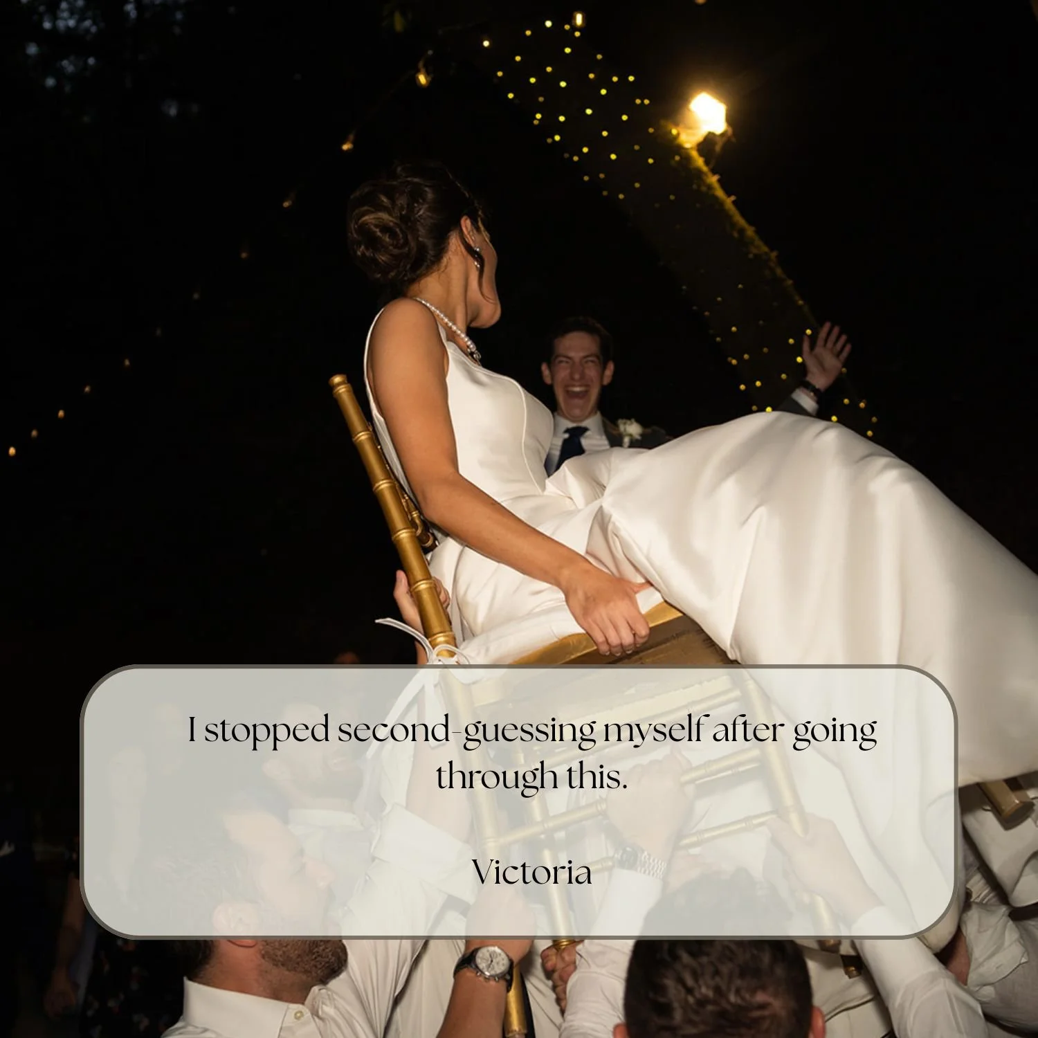 Bride and groom being lifted during a wedding celebration, with the groom smiling and the bride looking to the side, surrounded by party guests in a dark setting with string lights overhead.