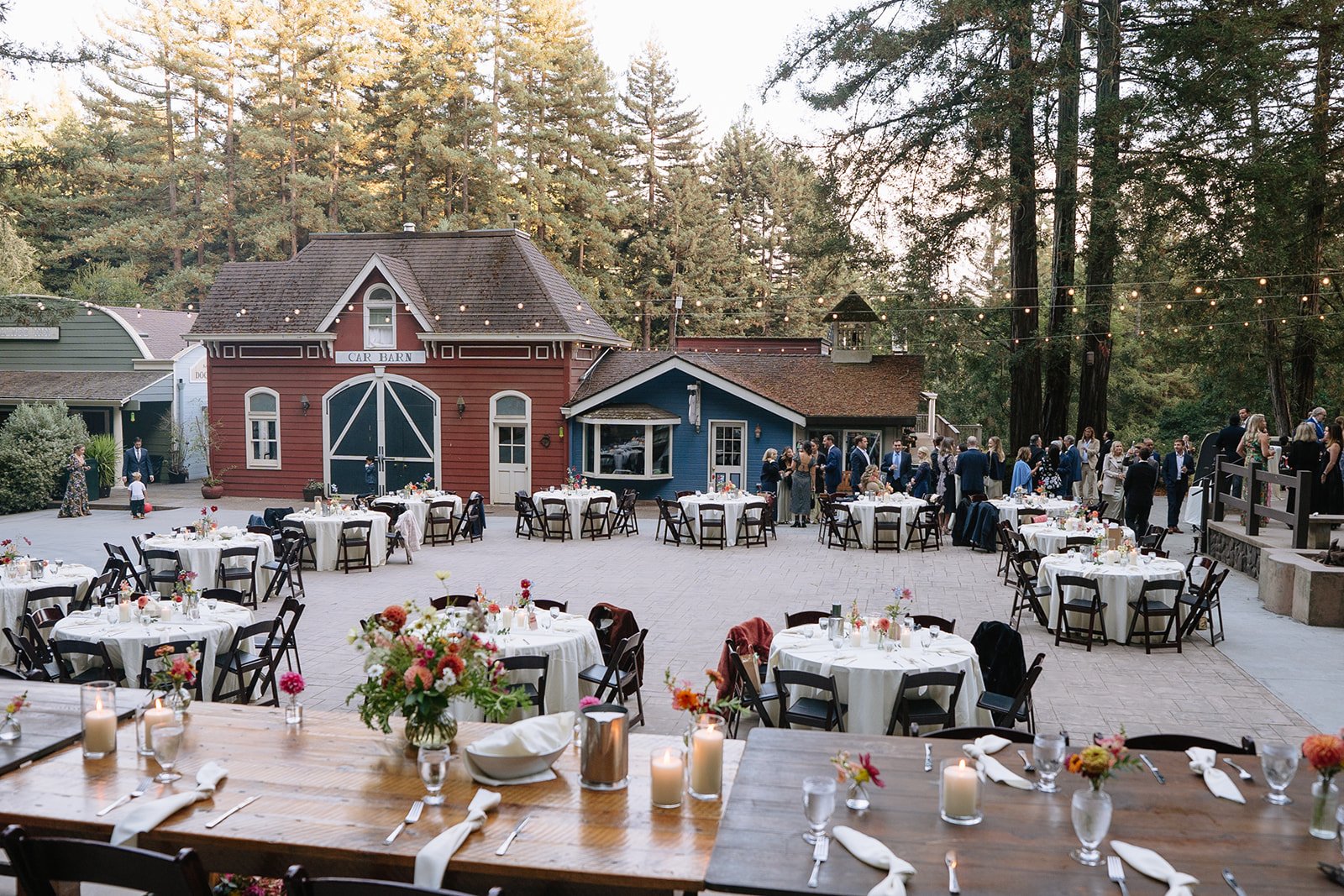 Outdoor wedding reception setup with round and rectangular tables decorated with flowers and candles, on a stone-paved open area with a colorful barn and pine trees in the background, during evening.