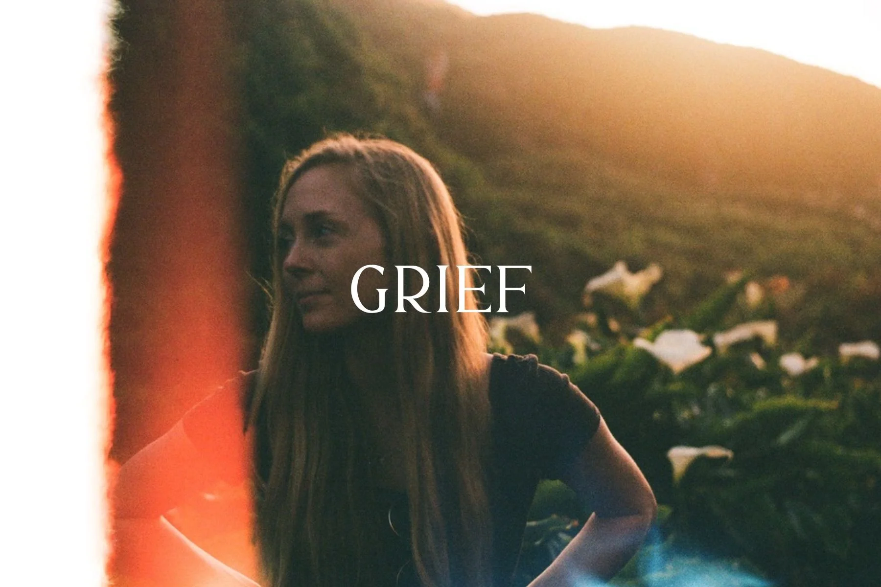A young woman standing outdoors at sunset or sunrise, with a hillside and wildflowers in the background, exhibiting a pensive or sad expression, with the word 'GRIEF' overlaid on the image.