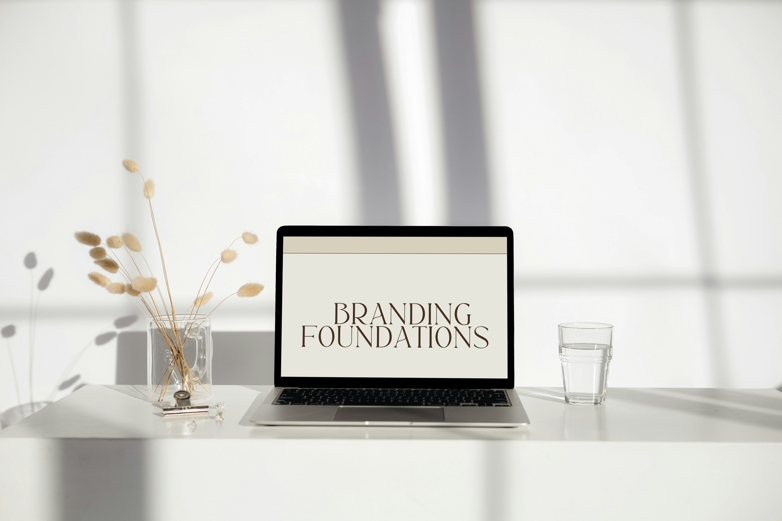 Laptop displaying the text 'Branding Foundations' on a white desk with a glass of water and a vase with dried flowers