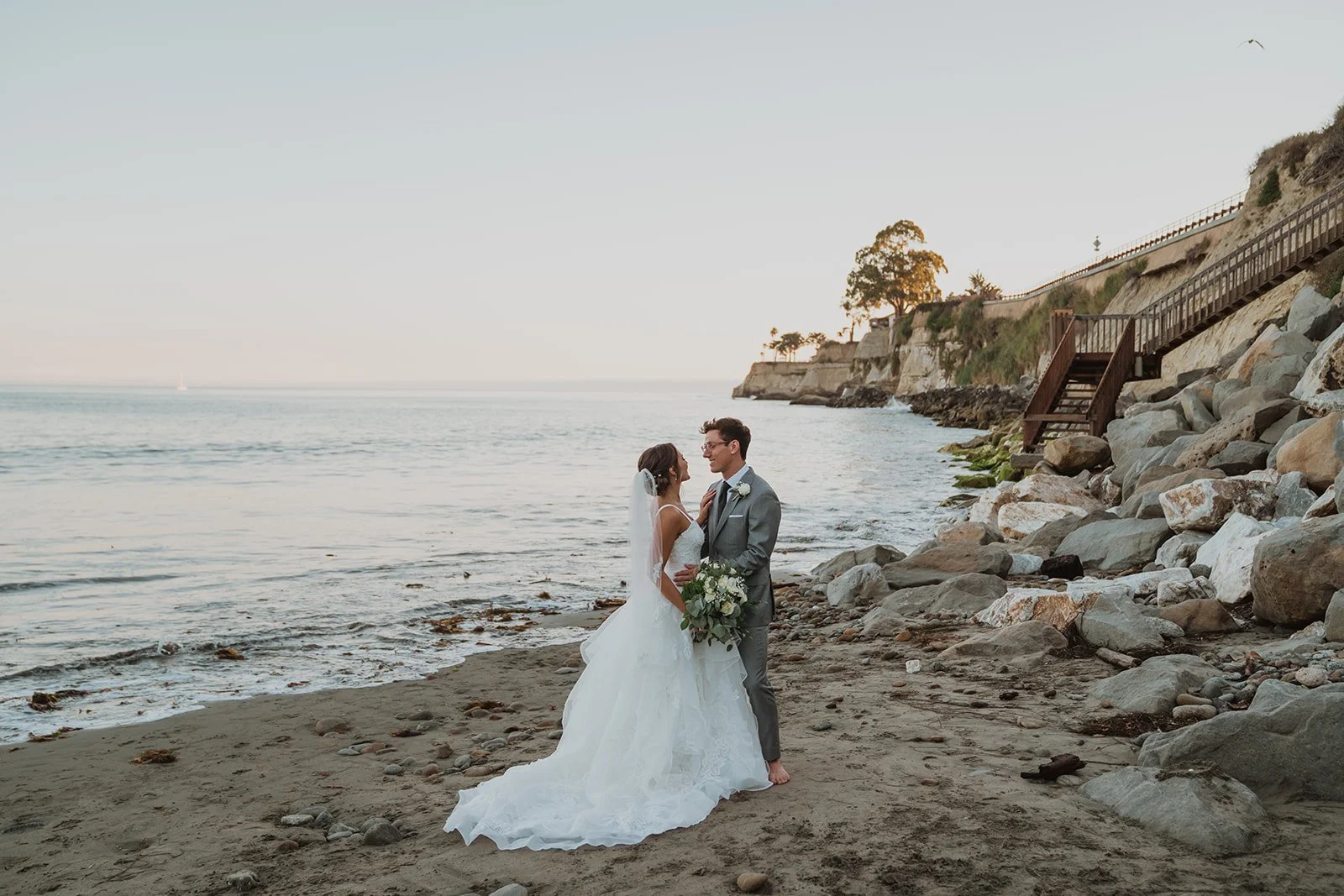 Bride and groom in wedding attire standing on sandy beach near rocky shoreline, holding bouquet, with cliffs and stairs in the background during sunset.