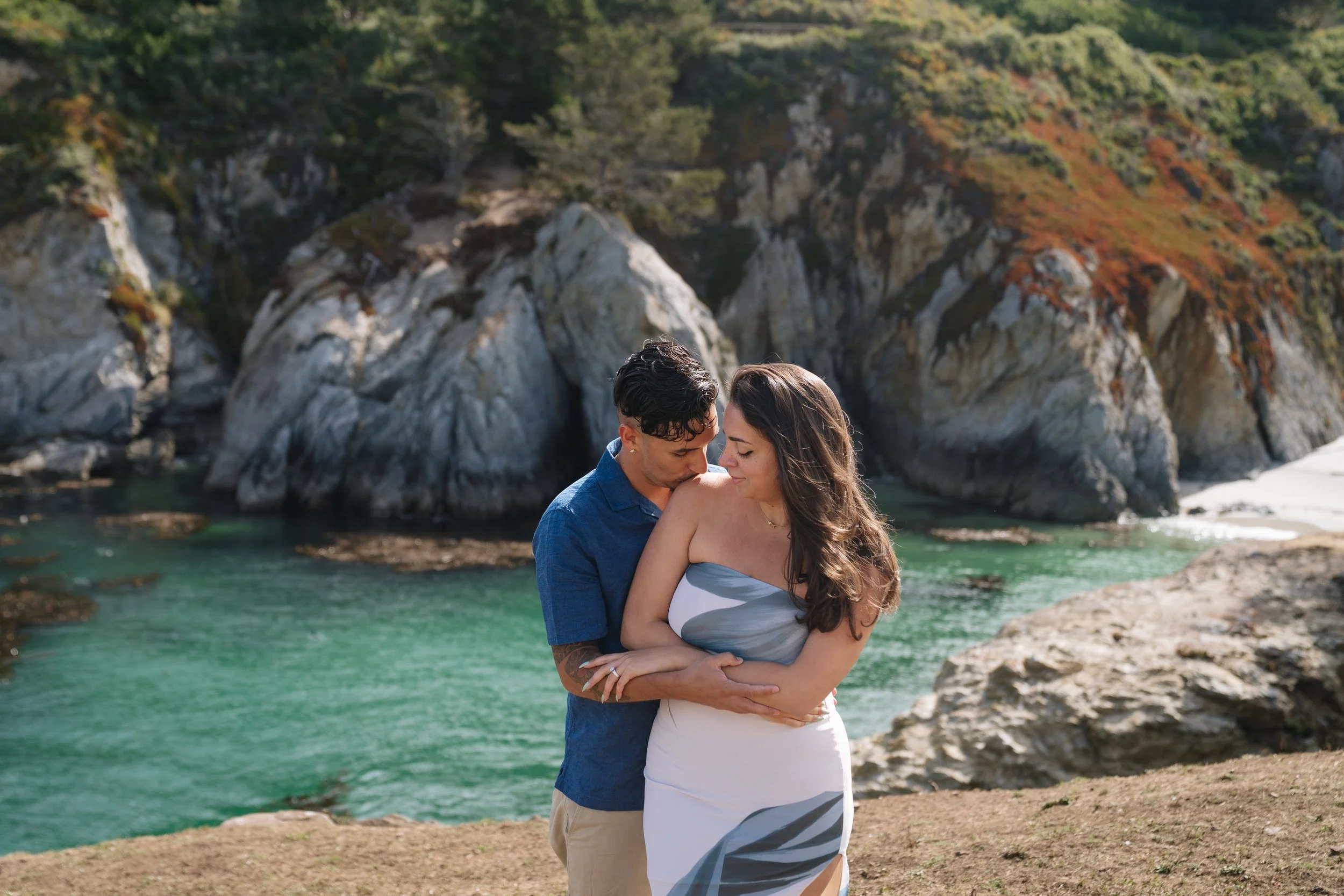A couple embracing by the water at a rocky, forested coastline.