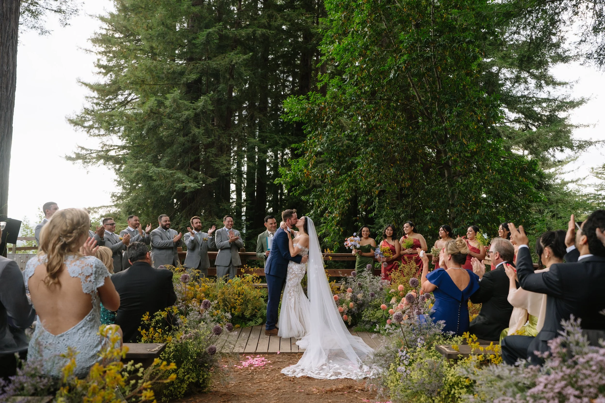 A wedding ceremony taking place outdoors with a couple kissing in front of guests, surrounded by flowers and tall trees in the background.