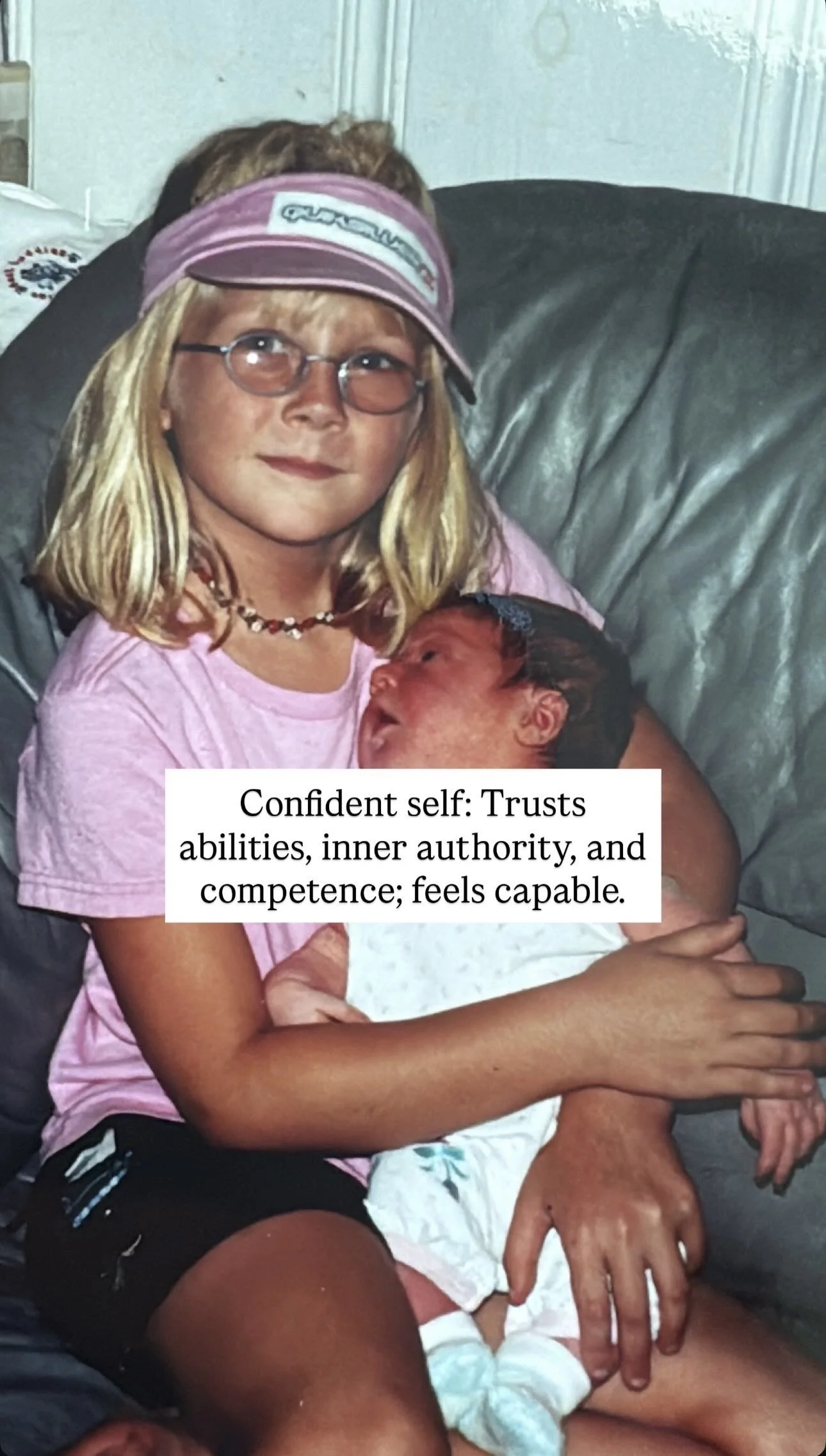 A young girl wearing glasses, a pink visor, and a pink T-shirt holds a newborn baby, sitting on a black leather couch. The girl has blond hair and a necklace, and the baby has dark hair and is dressed in white. The girl appears confident and caring.