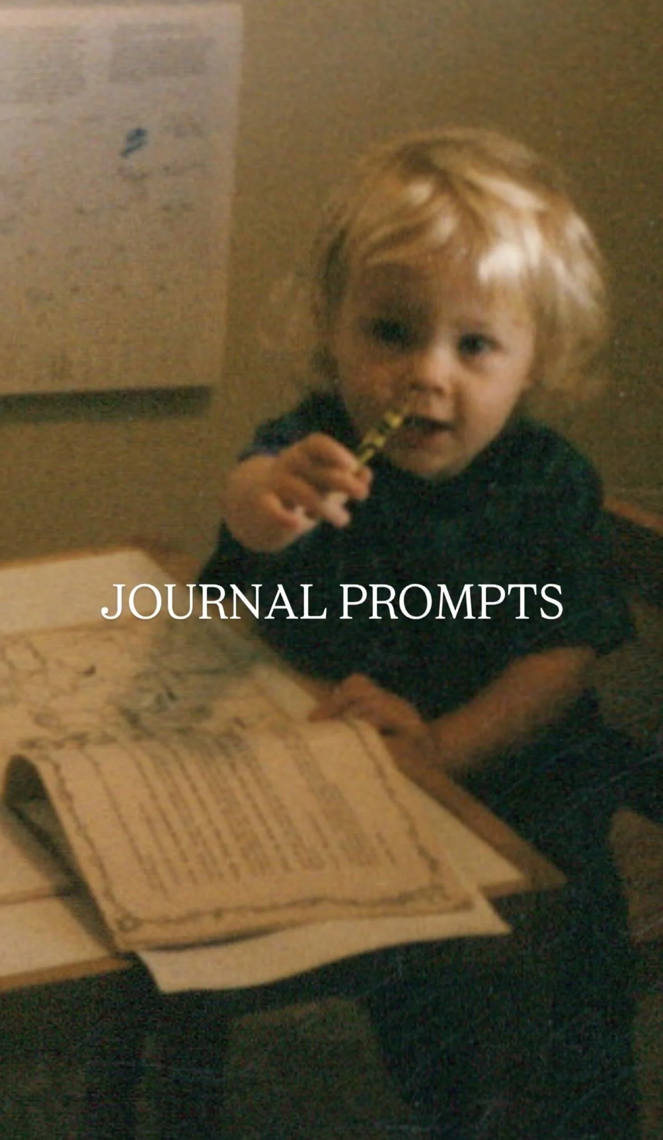 A young child with blonde hair sitting at a table with an open book, holding a yellow and black striped straw, with the text 'JOURNAL PROMPTS' overlaid.
