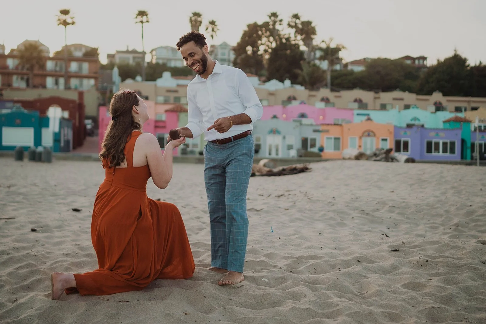 A man proposing marriage to a woman on the beach with colorful houses in the background.