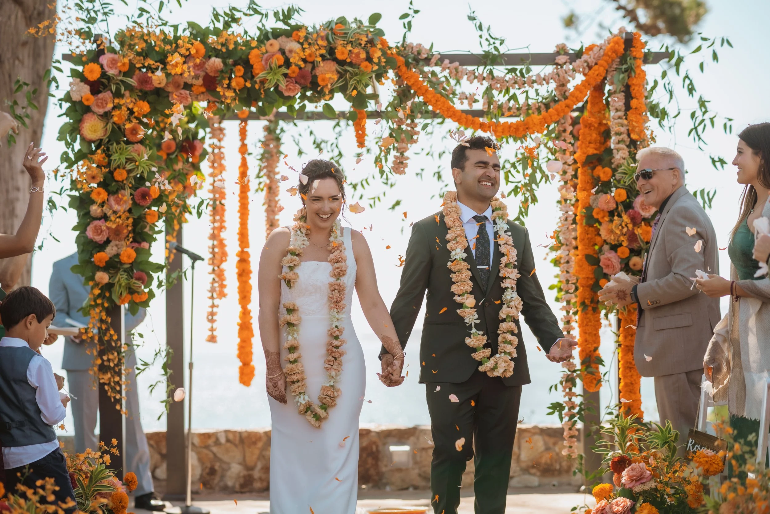 A wedding ceremony with a bride and groom holding hands, smiling, under a decorated floral arch with oranges, pinks, and greens. Guests are celebrating around them, some throwing flower petals.