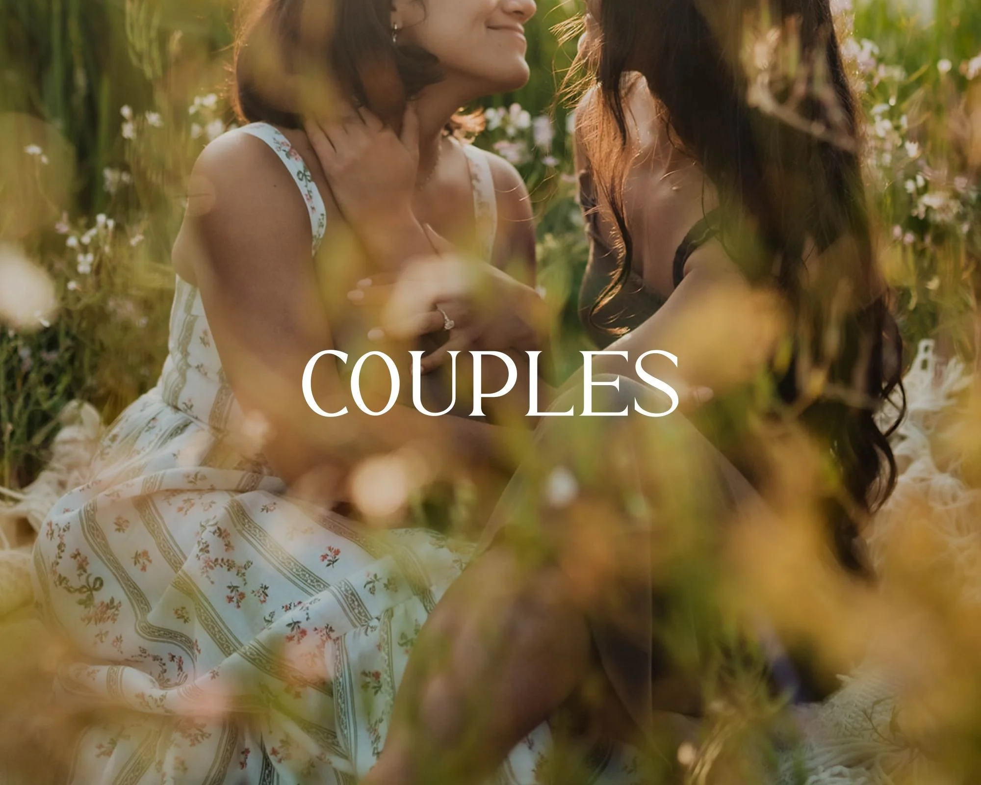 Two women sitting close together in a field of yellow flowers, facing each other affectionately. One woman has shoulder-length light brown hair and wears a sleeveless white dress with colorful patterns. The other woman has long dark hair and wears a dark dress. The scene appears warm and romantic, with the word 'COUPLES' overlayed in white text.