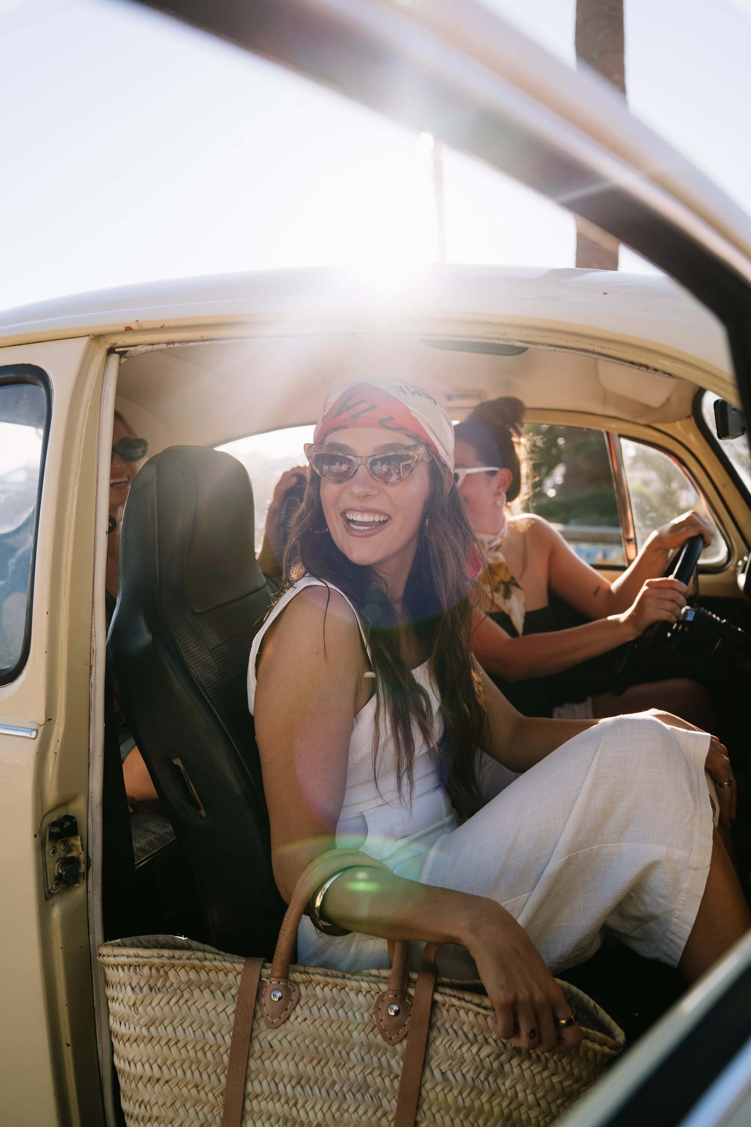 A group of women enjoying a car ride on a sunny day, with one woman smiling and wearing sunglasses and a bandana, sitting in the front seat of a vintage car.