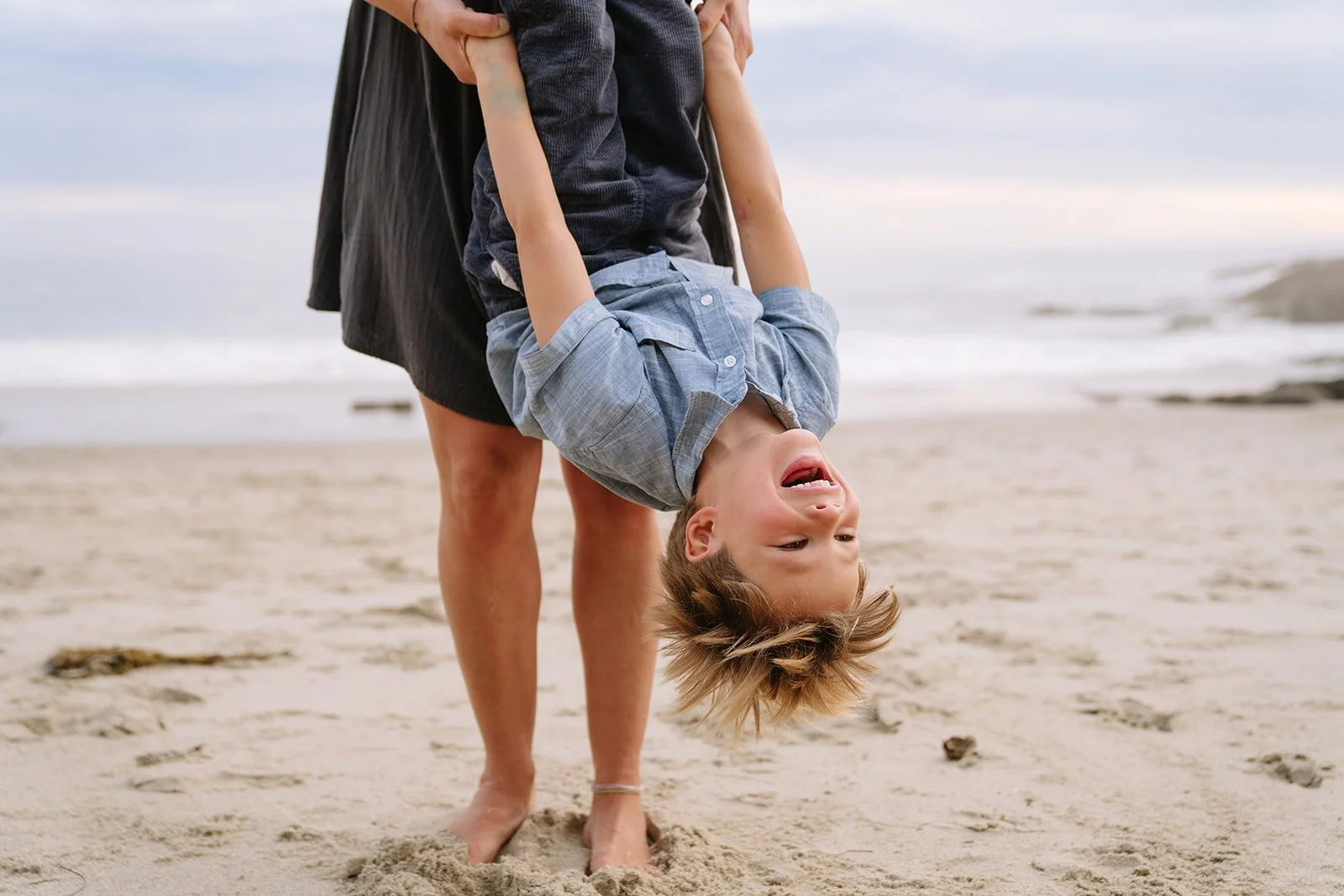 A person holding a laughing boy upside down on a sandy beach with the ocean and cloudy sky in the background.