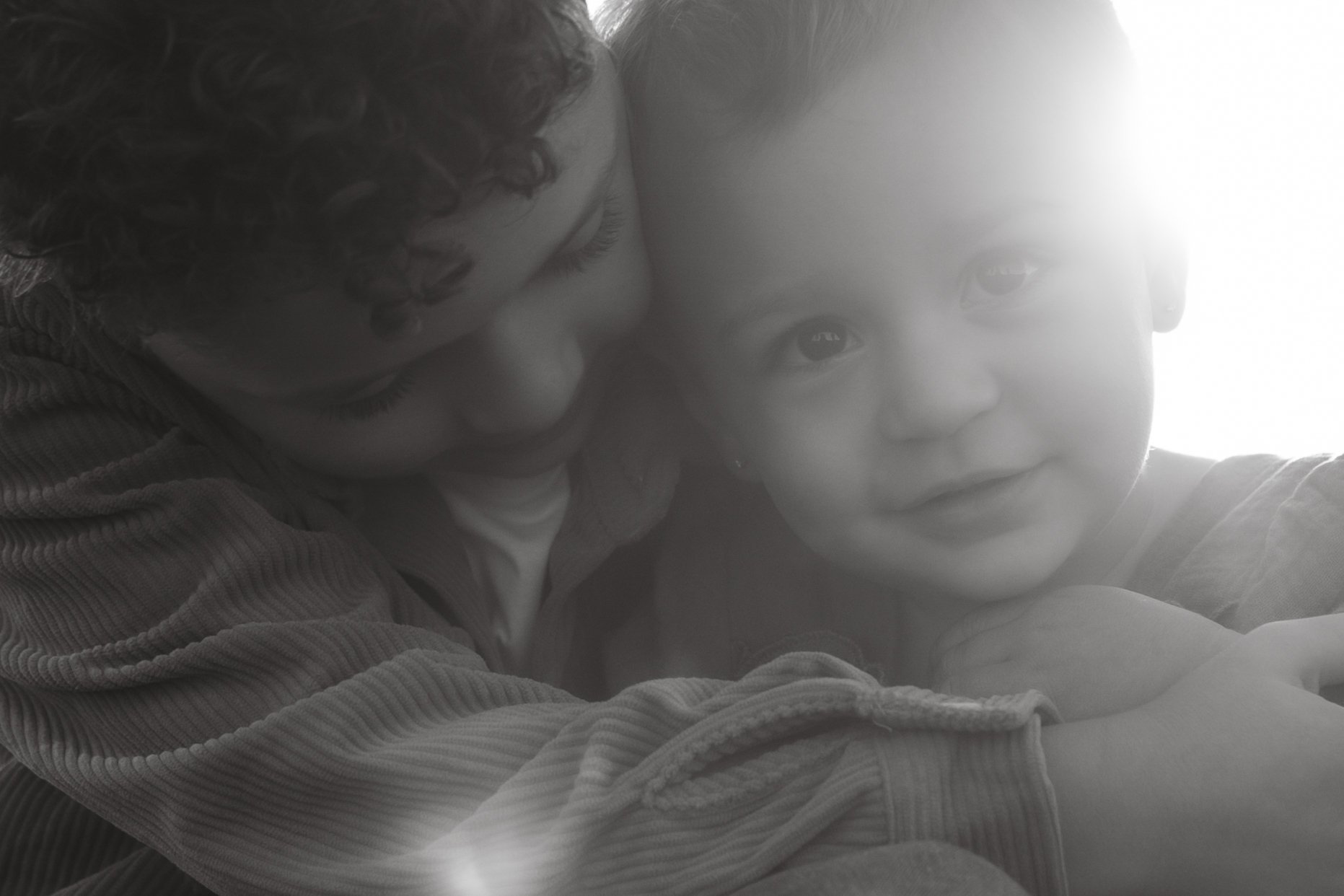 A woman and a young child close up, with the woman leaning in towards the child, both looking at the camera, with soft lighting behind them.