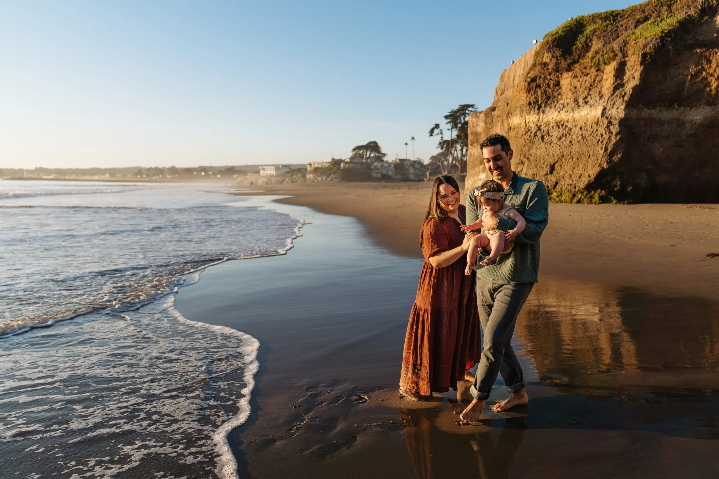 A family of three walking barefoot on the beach during sunset, with a cliff and trees in the background.