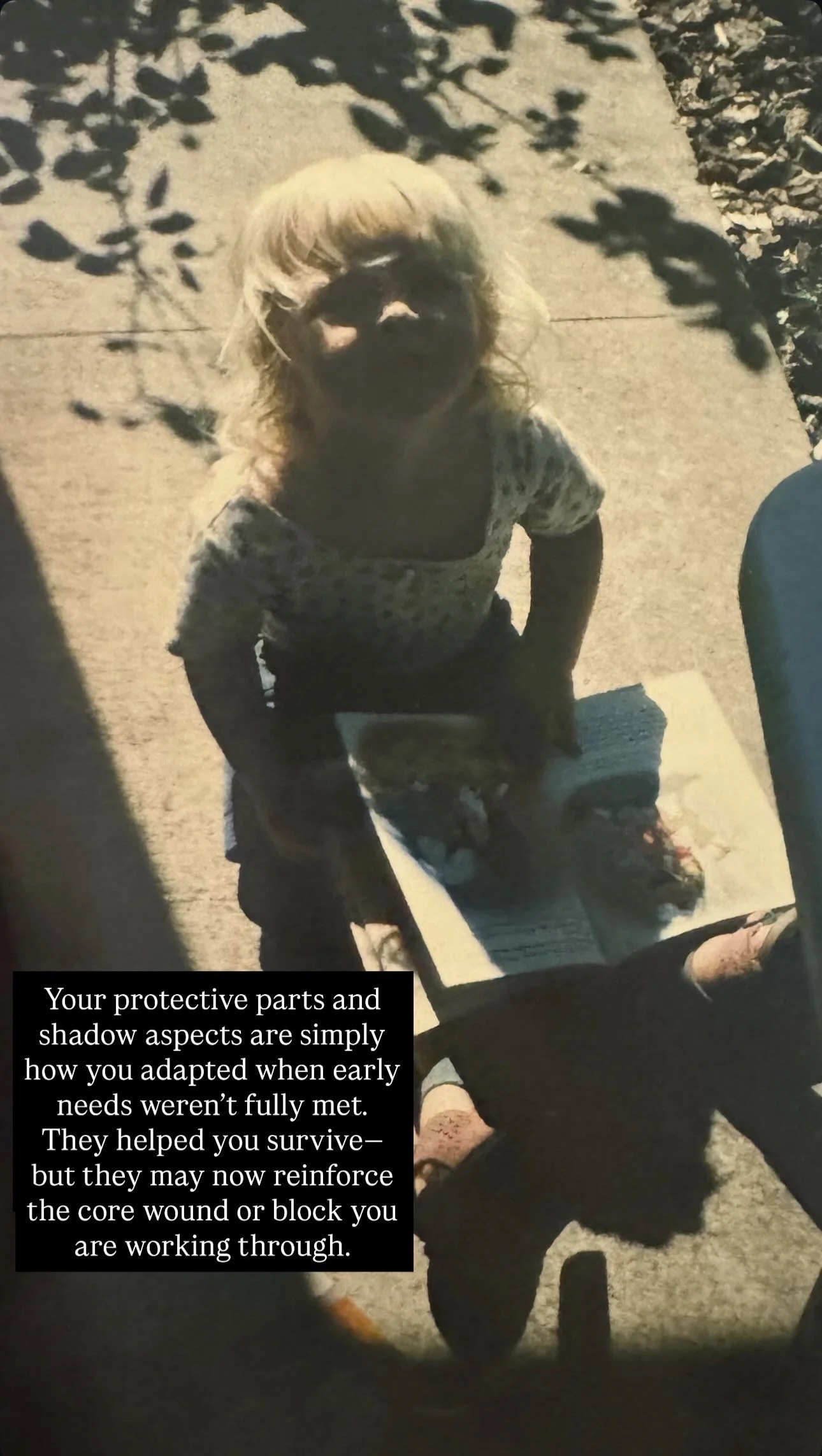 A young girl with blonde hair looking up at the camera with shadowed features, standing on a sidewalk next to a table or surface. There is a shadow of a tree's branches and leaves on the ground and on the girl. The girl appears to be standing over a 