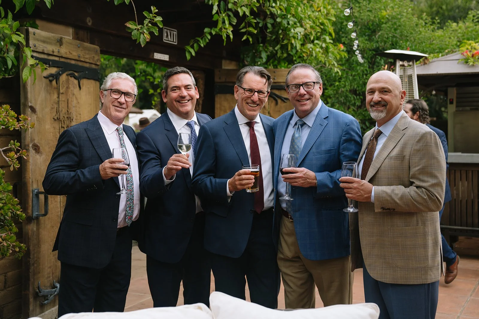 Six professionally dressed men standing close together at an outdoor event, smiling, and holding drinks, with greenery and string lights in the background.